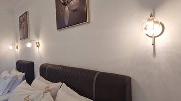 A hotel room with a bed, white pillows with embroidered text, a wall-mounted light fixture, and framed floral artwork.