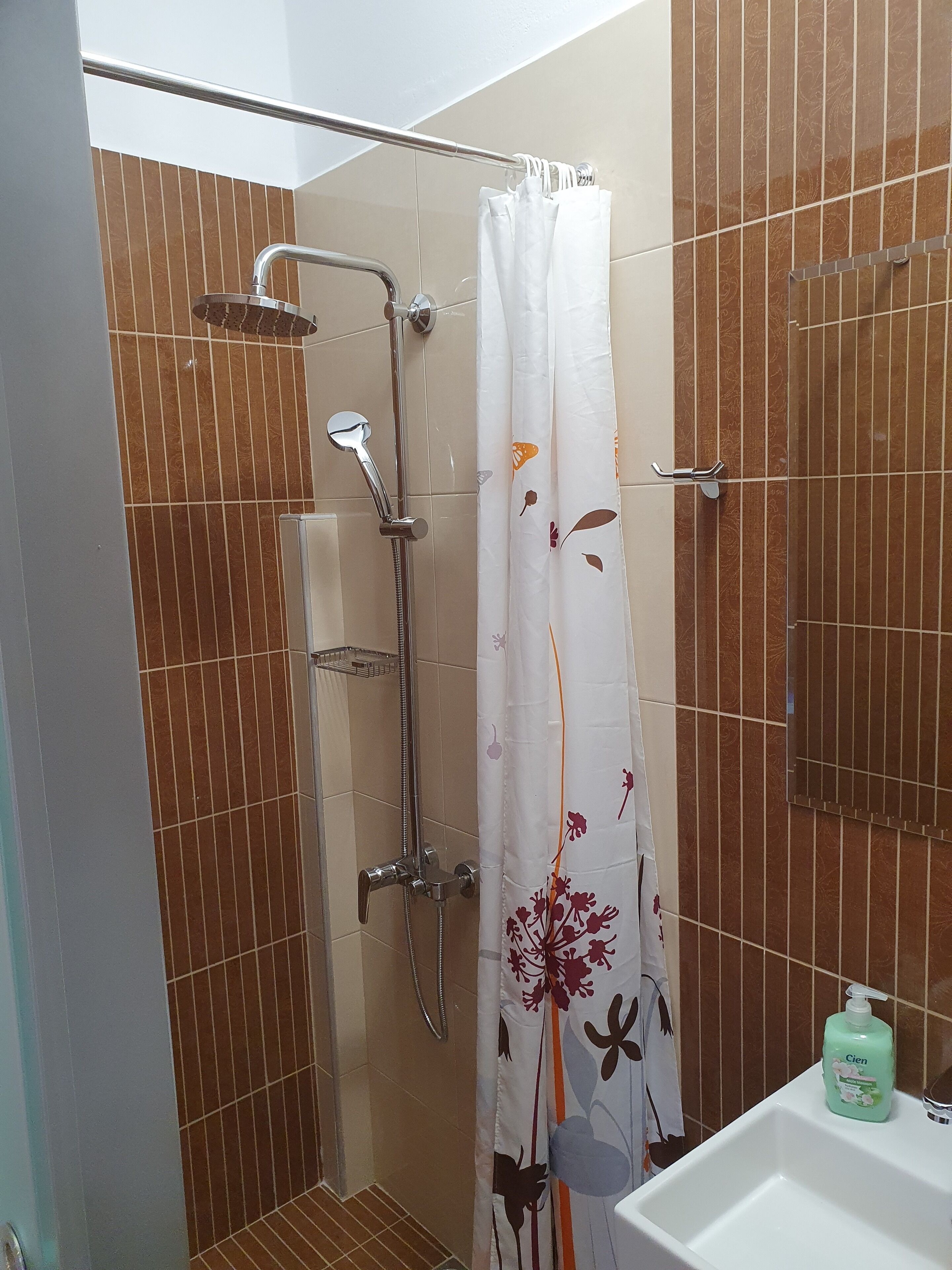 A shower with a rain showerhead, a handheld showerhead, and a handheld shower attachment. There is a white shower curtain with a floral pattern. A bottle of Cien soap is visible on the sink.