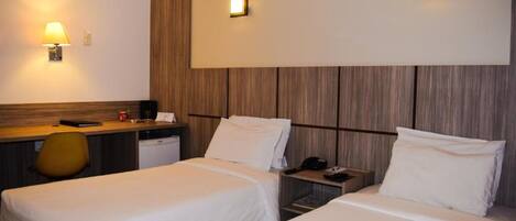 A hotel room with two single beds, a desk with a lamp, a chair, a small table, and a wall-mounted phone.