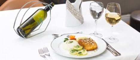 Une table à manger recouverte d’une nappe blanche, une bouteille de vin, deux verres à vin, une assiette avec de la nourriture et des couverts.