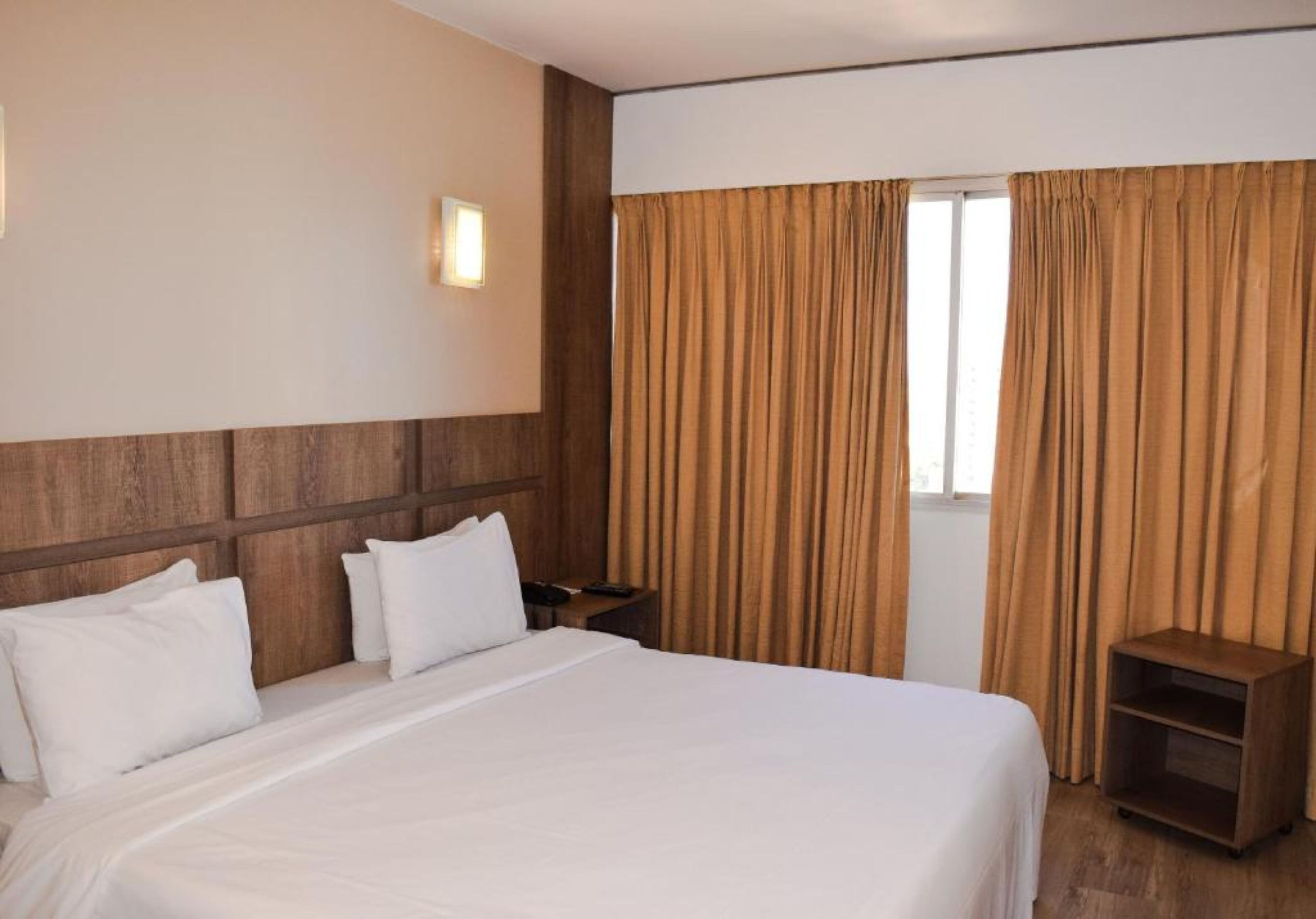 Ein Hotelzimmer mit einem großen Bett, einem Holzkopfstück, einem Nachttisch, einem kleinen Seitentisch und einem Fenster mit Vorhängen.