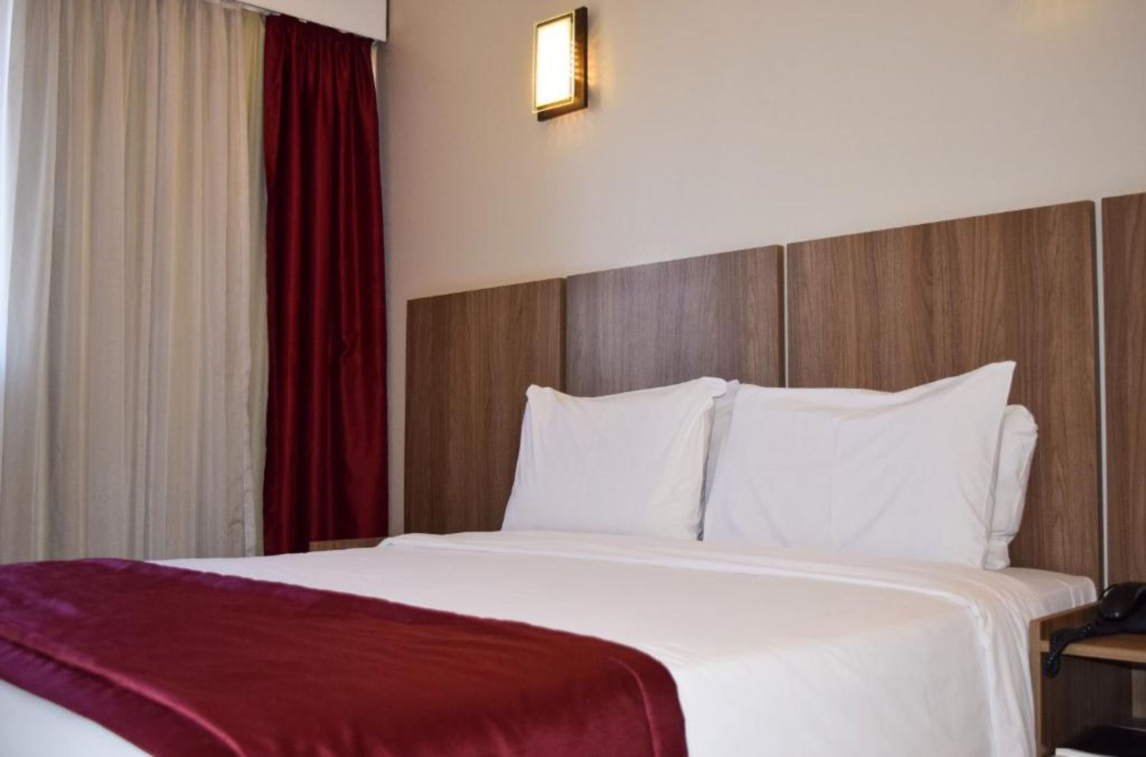 Una habitación de hotel con una cama grande, cabecera de madera, cortina roja y teléfono junto a la cama.