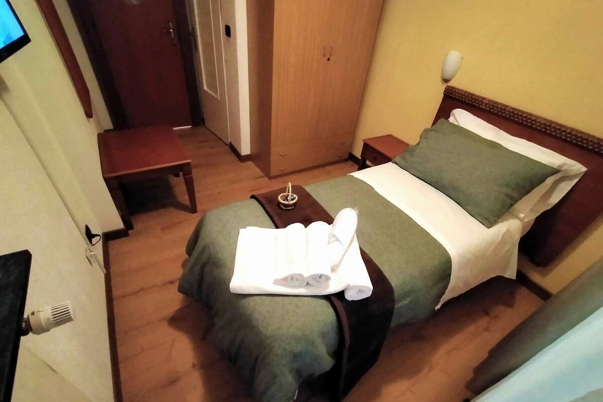 A hotel room with a bed, a bedside table, a nightstand, a small table, and a TV on the wall.