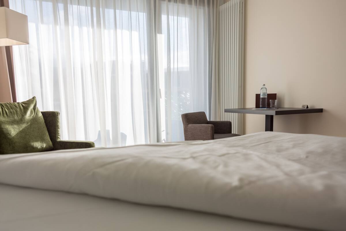 A hotel room with a large bed, a chair, a small table with a bottle and glass, and sheer curtains.