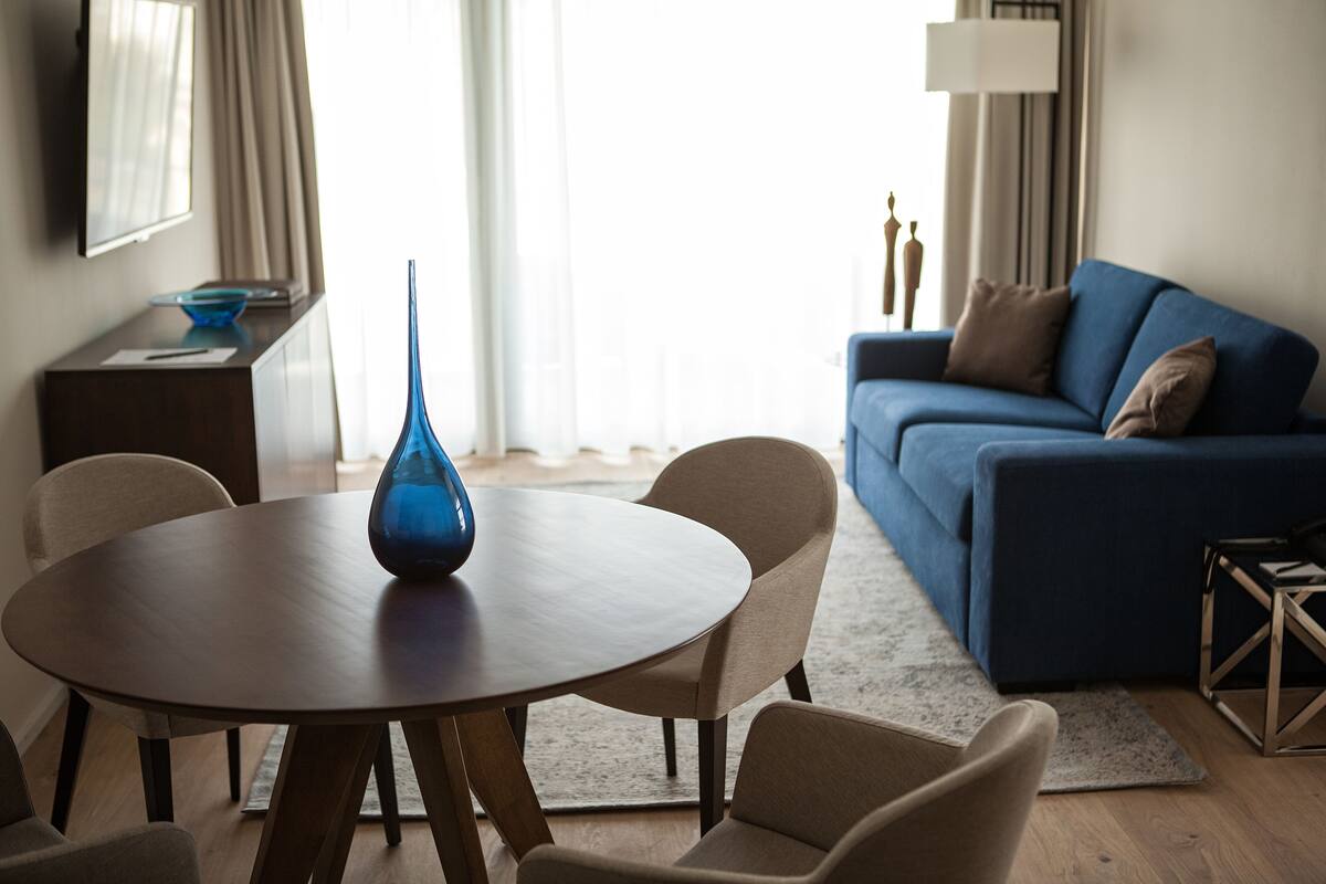 A modern living room with a round wooden table, beige chairs, a blue vase, a blue sofa, and a lamp.