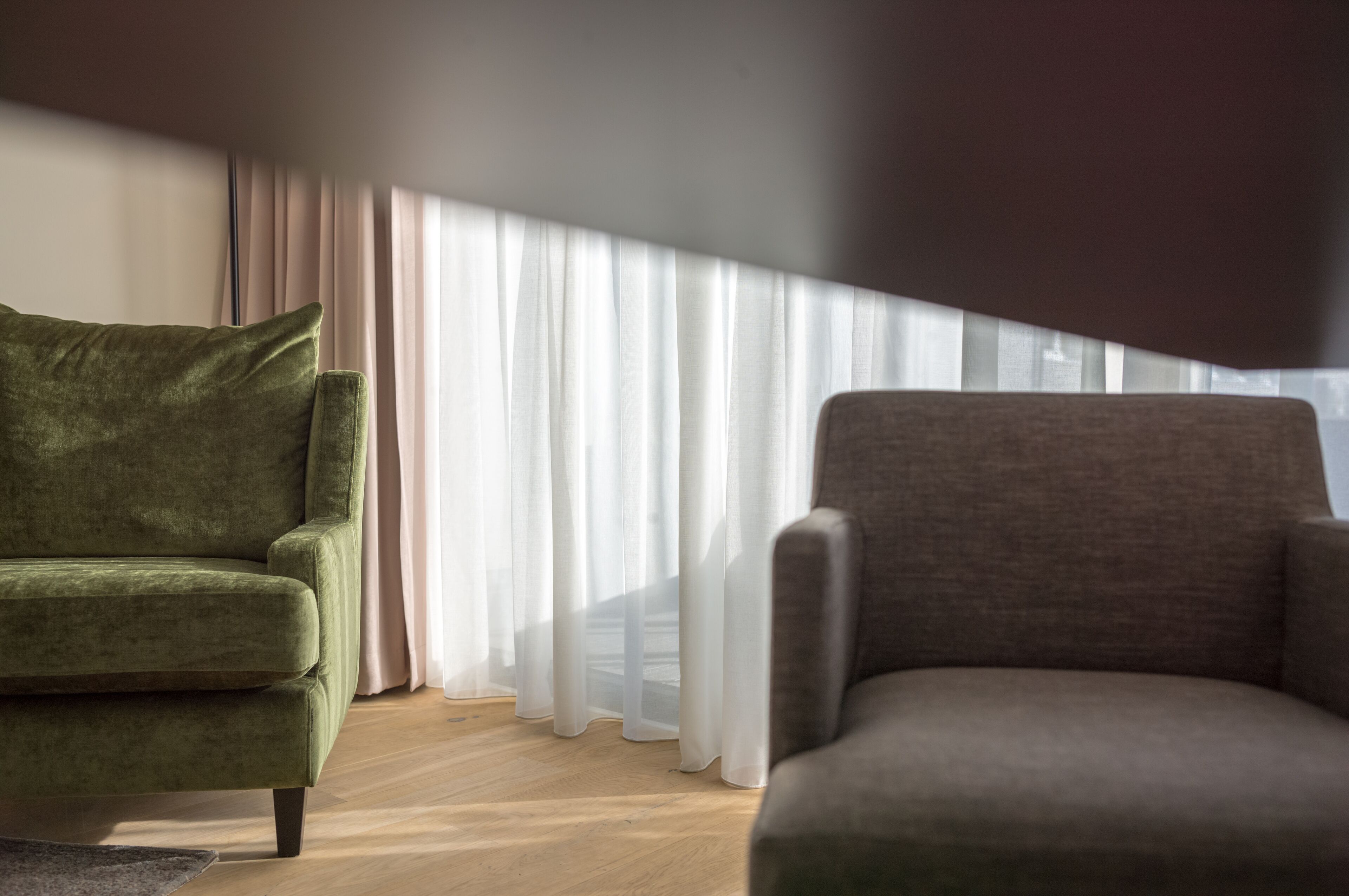 A room with a green armchair and a grey armchair, a large window with sheer curtains, and a wooden floor.
