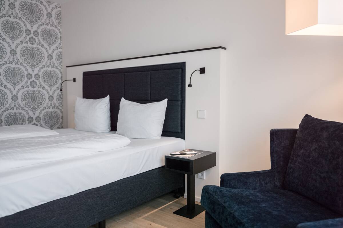A modern hotel room with a large bed, a dark headboard, a side table, and a dark armchair.