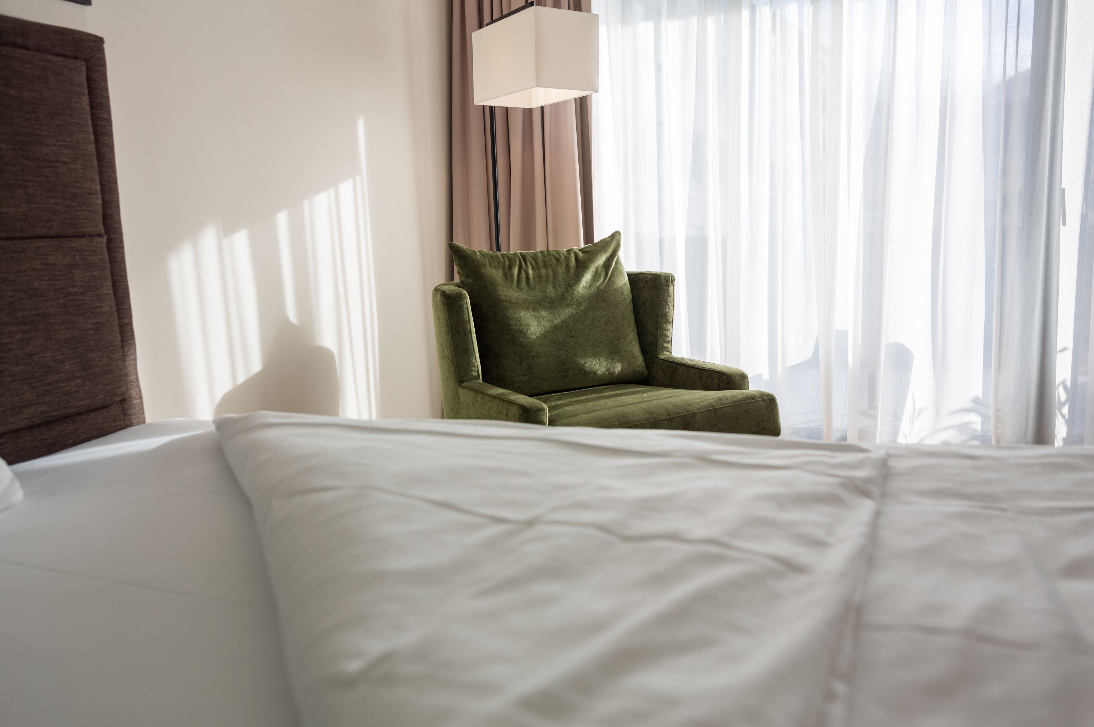 Une chambre d’hôtel avec un lit, un fauteuil vert, une lampe et des rideaux en voile.