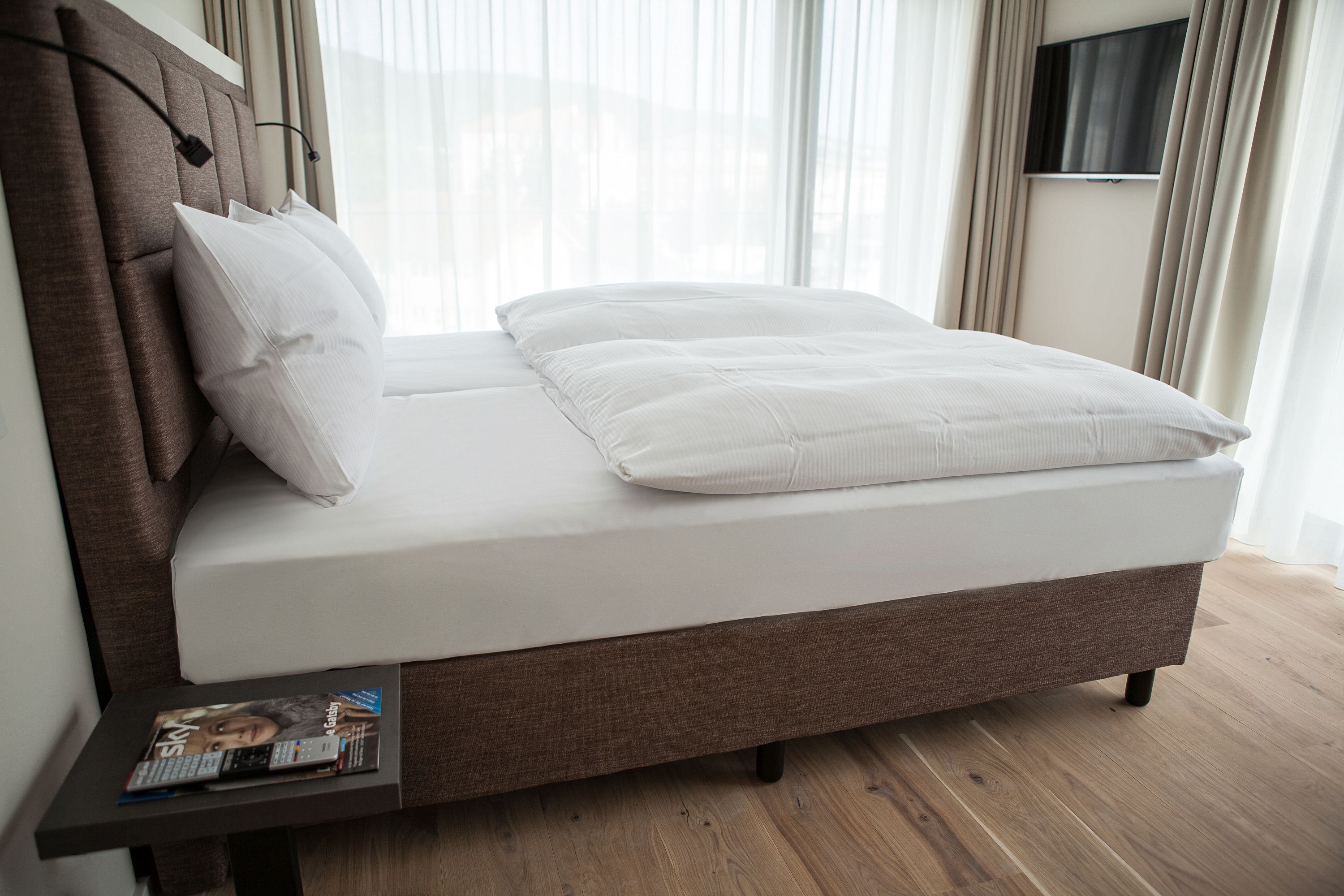 Una cama bien tendida con cabecera, una mesita de noche con una revista, y un televisor montado en la pared.