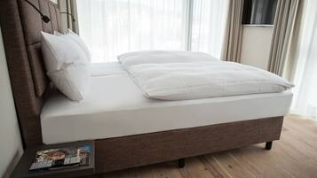 Una cama bien tendida con cabecera, una mesita de noche con una revista, y un televisor montado en la pared.