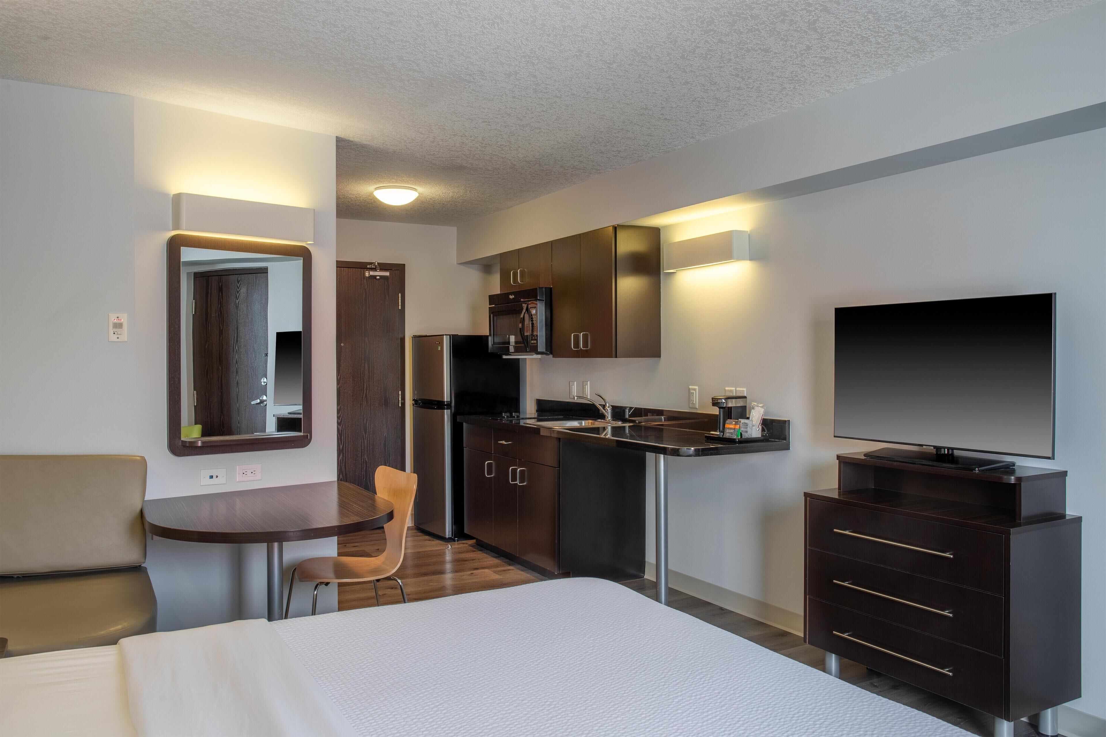 A compact hotel room with a bed, a small dining table, a chair, a kitchenette with a microwave, refrigerator, and a TV.