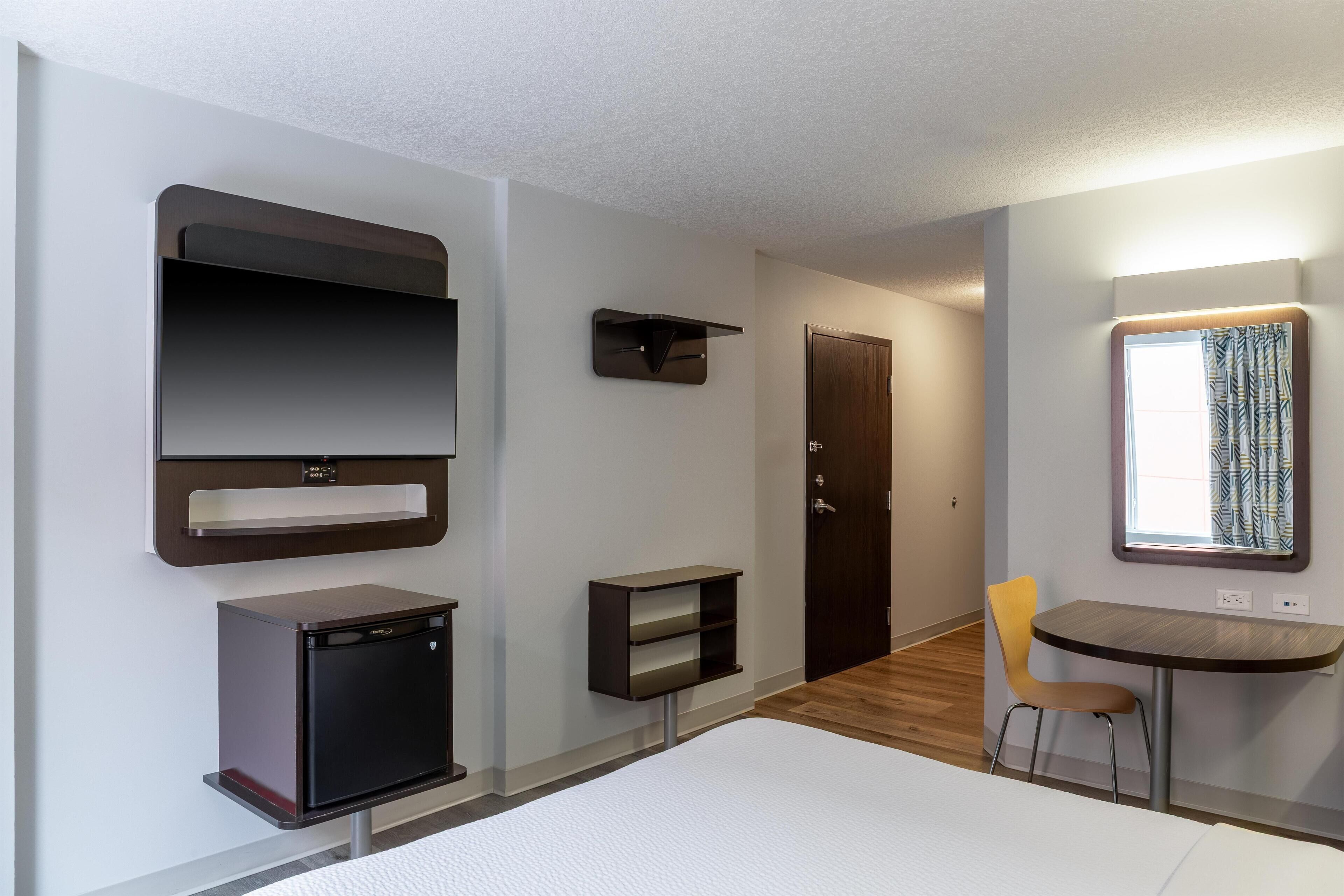 Habitación de hotel compacta con cama, pequeño escritorio con silla, televisor integrado, refrigerador minibar, armario y ventana con cortinas.