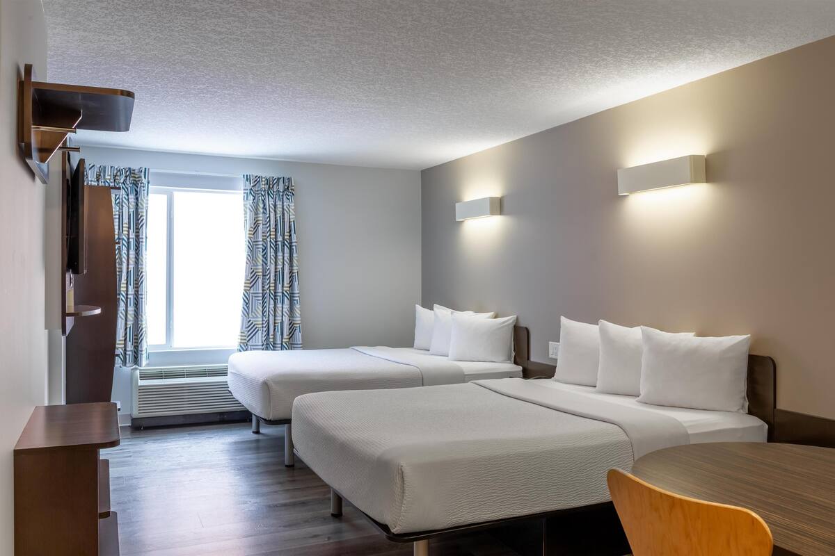Une chambre d’hôtel avec deux lits, un bureau et une chaise. Il y a une fenêtre avec des rideaux à motifs, un téléviseur fixé au mur et un luminaire fixé au mur.