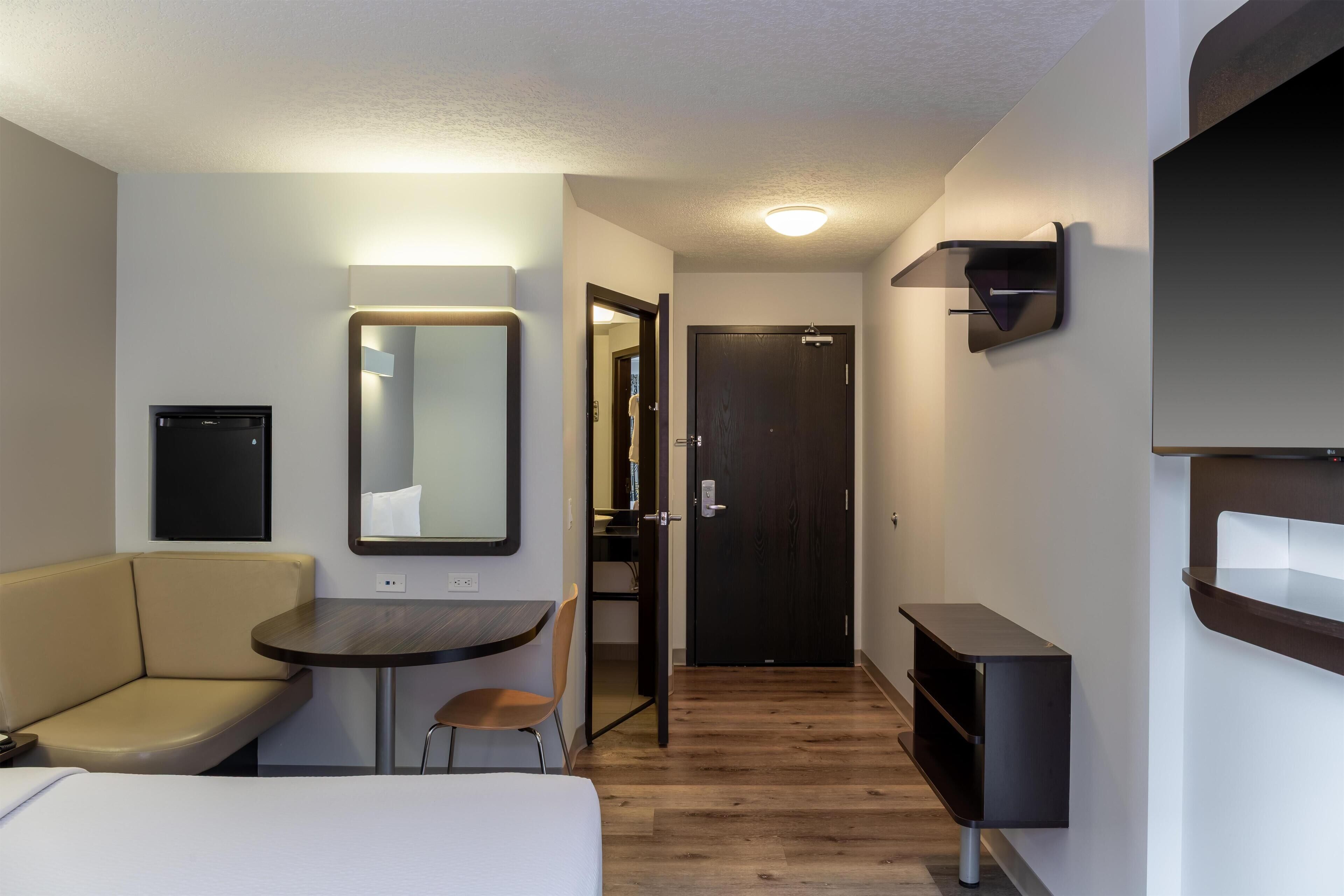 A compact hotel room with a bed, a small table, a chair, a sofa, a mirror, a door, and a wall-mounted shelf.