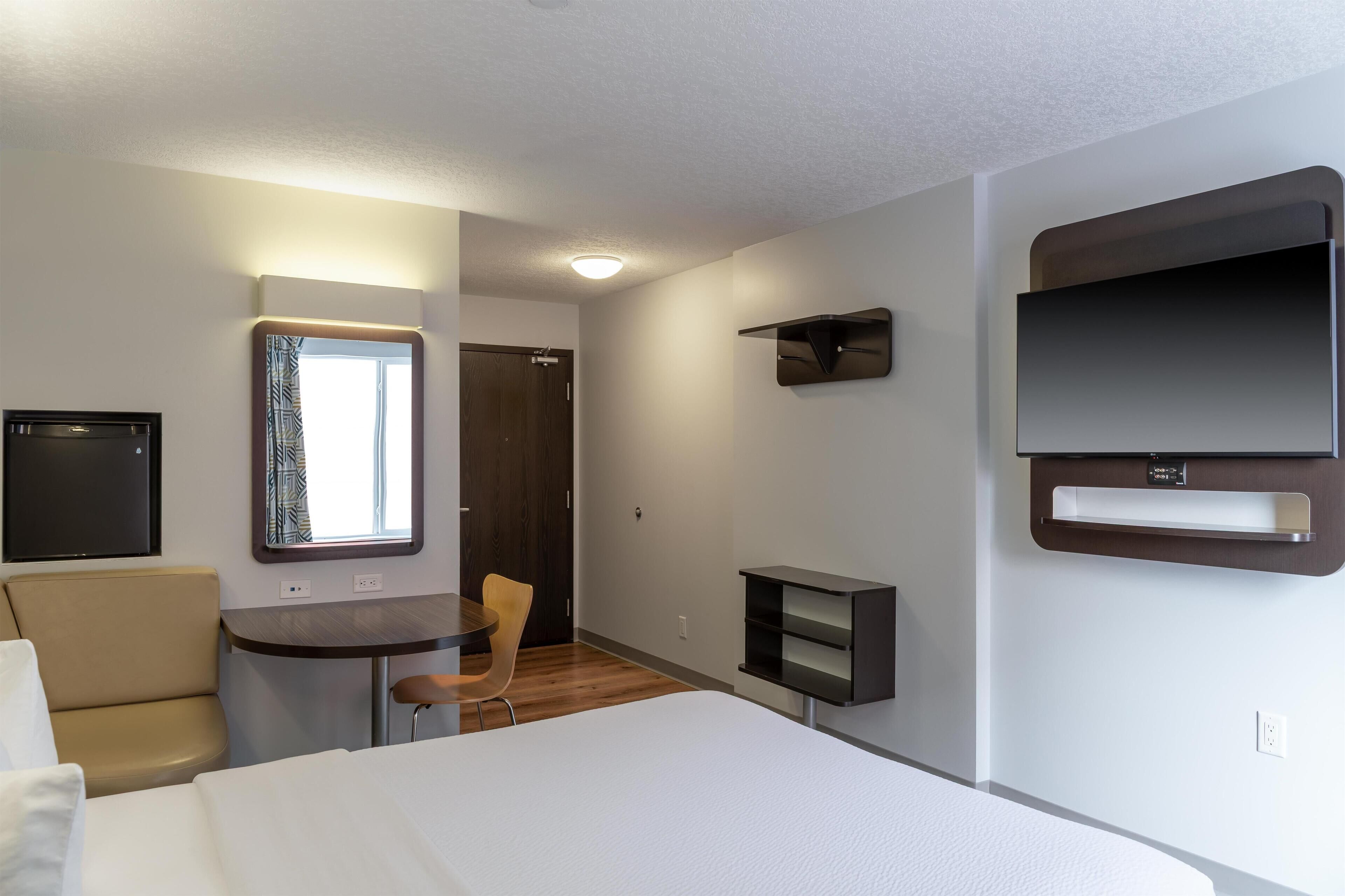 Una habitación de hotel moderna con una cama, un escritorio, una silla, un televisor montado en la pared y un ventanal con cortinas.