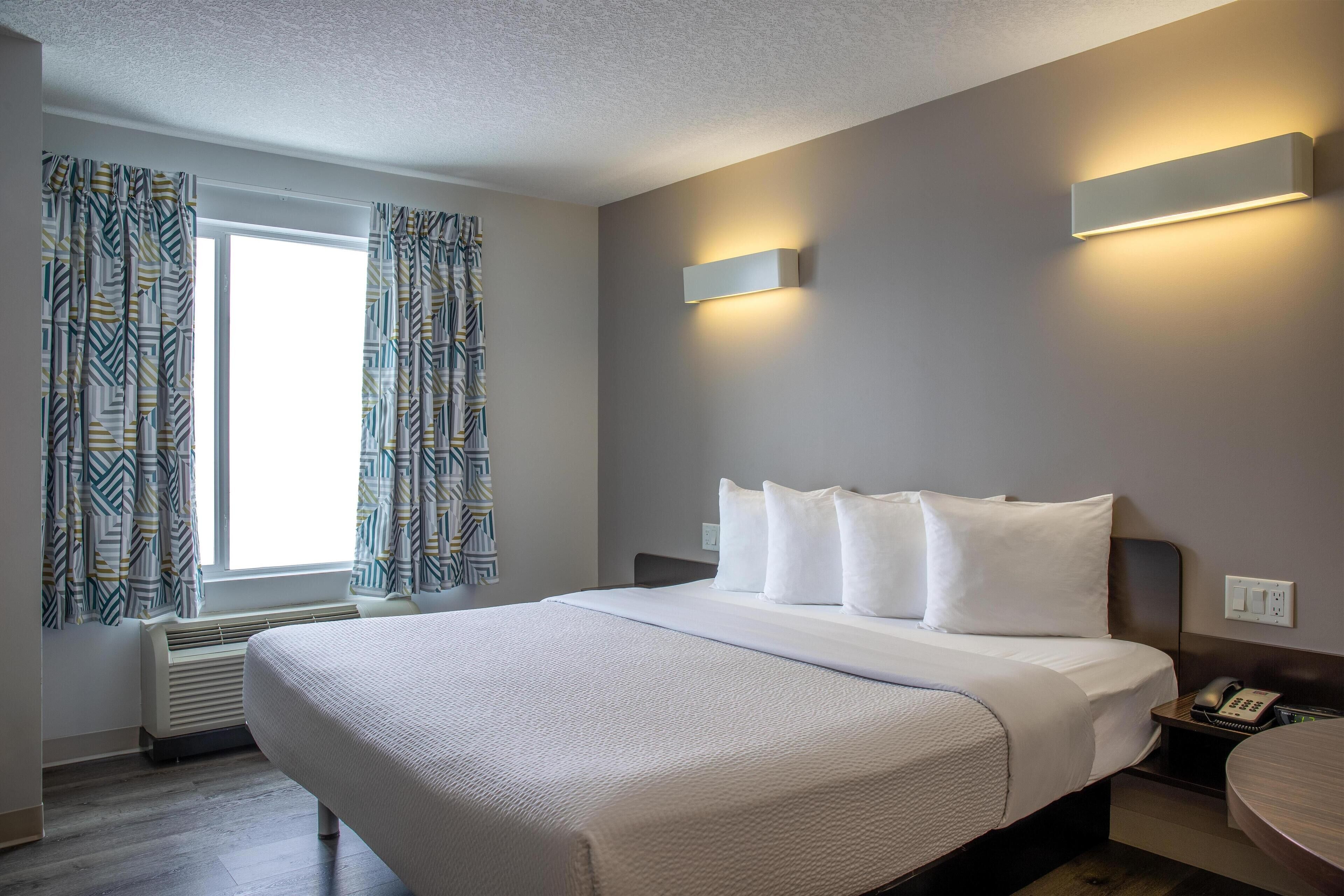 Een hotelkamer met een groot bed, een nachtkastje met een lamp, een raam met gordijnen met een patroon, en een wandlamp.