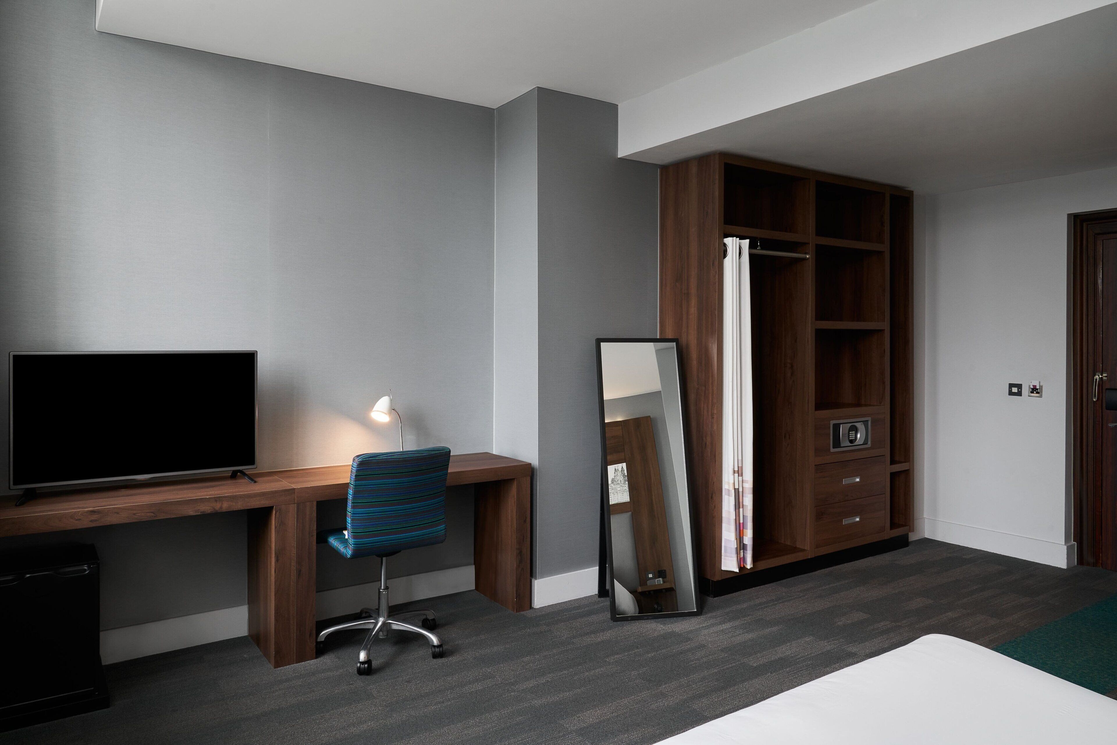 Una habitación moderna con un escritorio de madera, una silla, televisor de pantalla plana, un espejo y un armario.