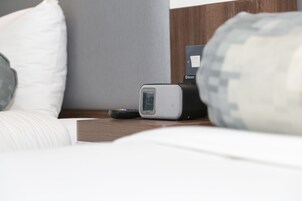 A bed with a white pillow, a remote control, and a digital clock on a bedside table.