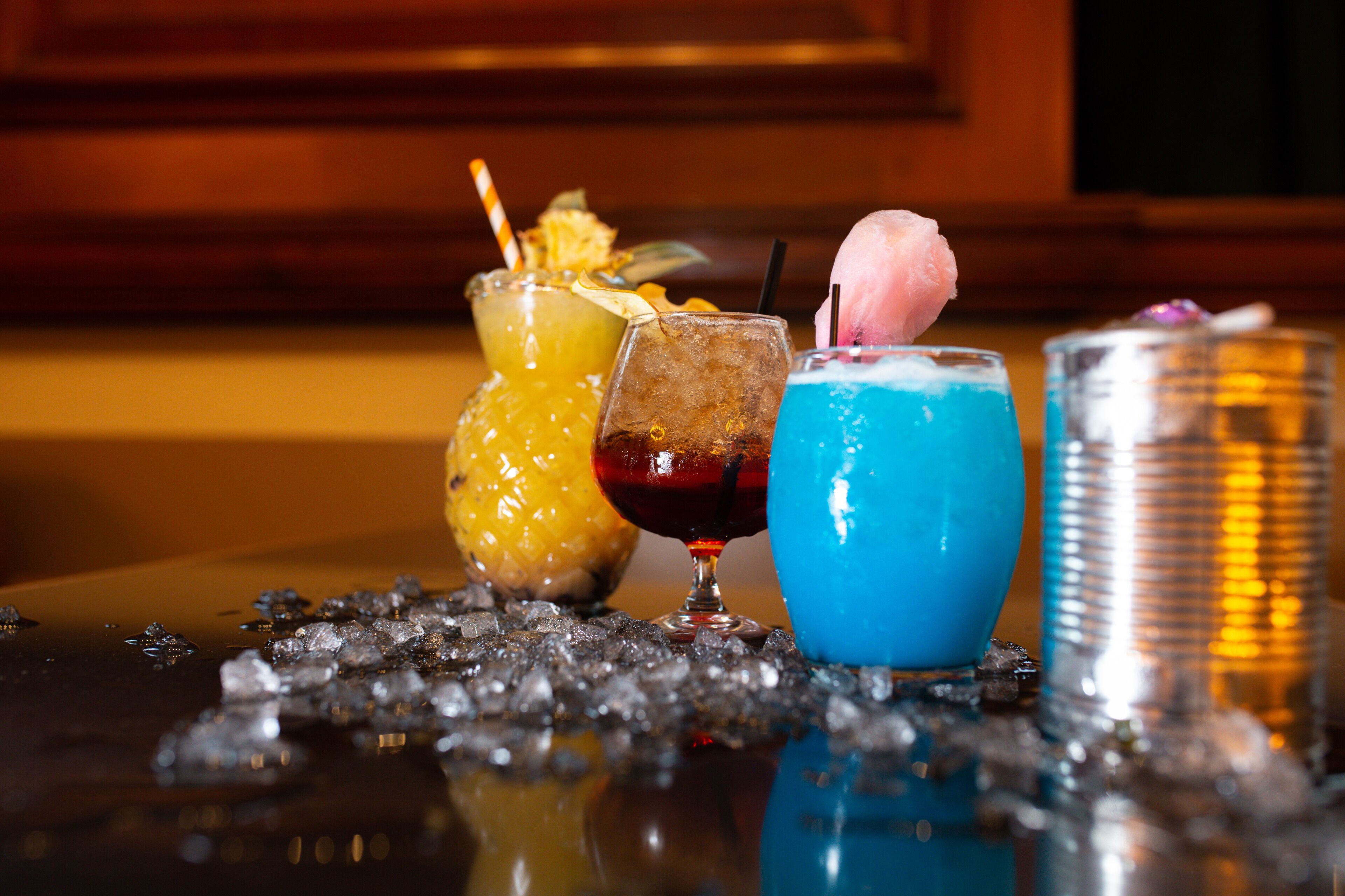 Trois cocktails sont posés sur le comptoir d’un bar, accompagnés de glaçons, de décorations et d’un grand verre contenant une boisson bleue.