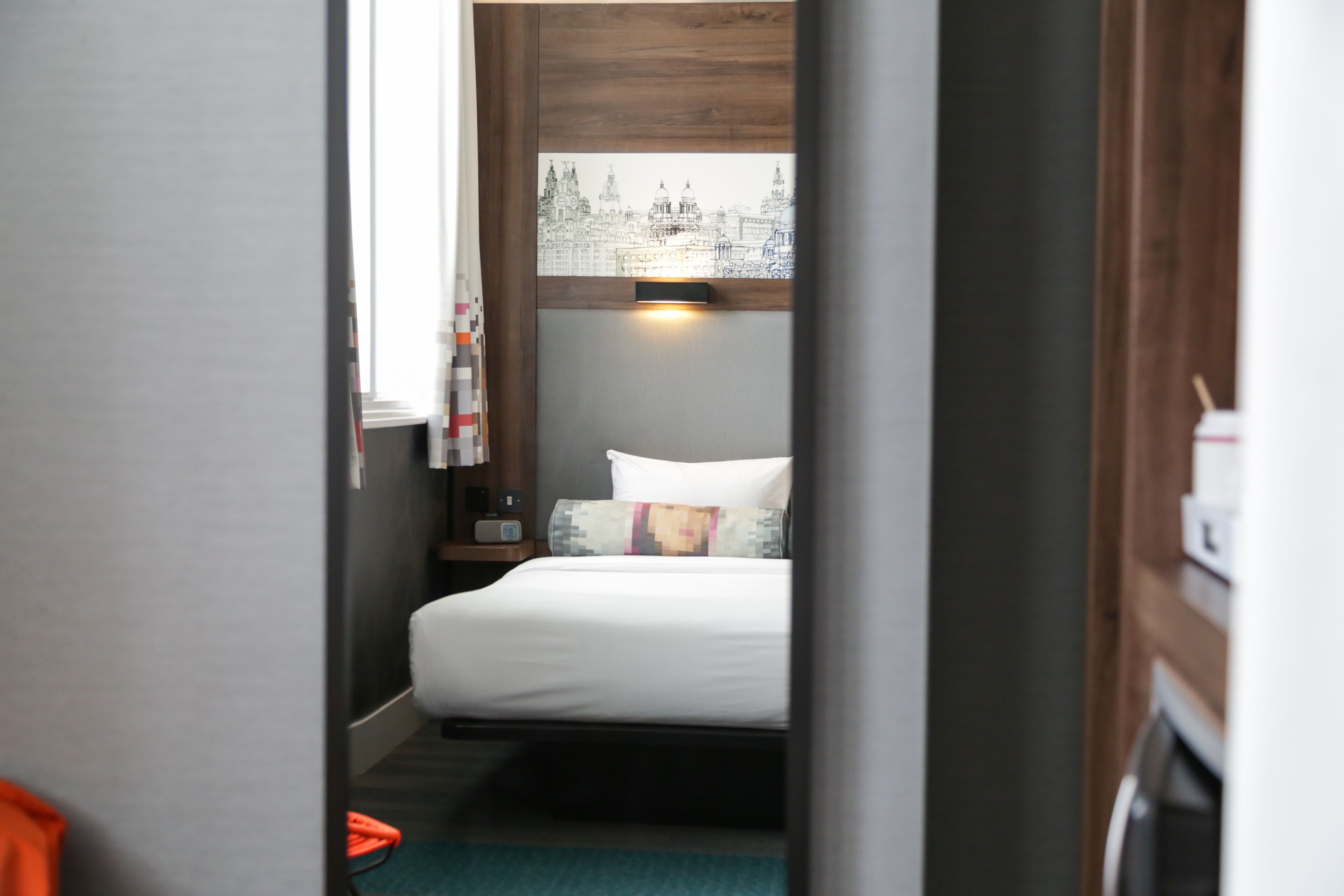 A narrow hallway leading to a bed with a cityscape view, a bedside table, and a nightstand with a lamp.