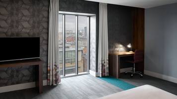 Une chambre d’hôtel avec une grande fenêtre, un bureau avec une chaise, une télévision et un lit.