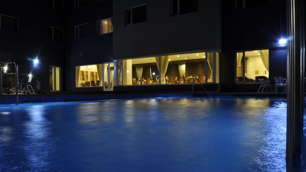 Vista nocturna de un hotel con piscina al aire libre, zona de comedor iluminada y ventanas visibles.