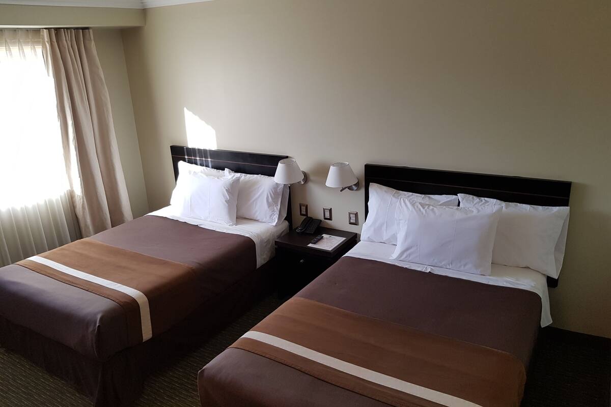 Habitación de hotel con dos camas, cada una con sábanas blancas y cabeceras marrones. Hay una mesita de noche con una lámpara y un teléfono entre las camas.
