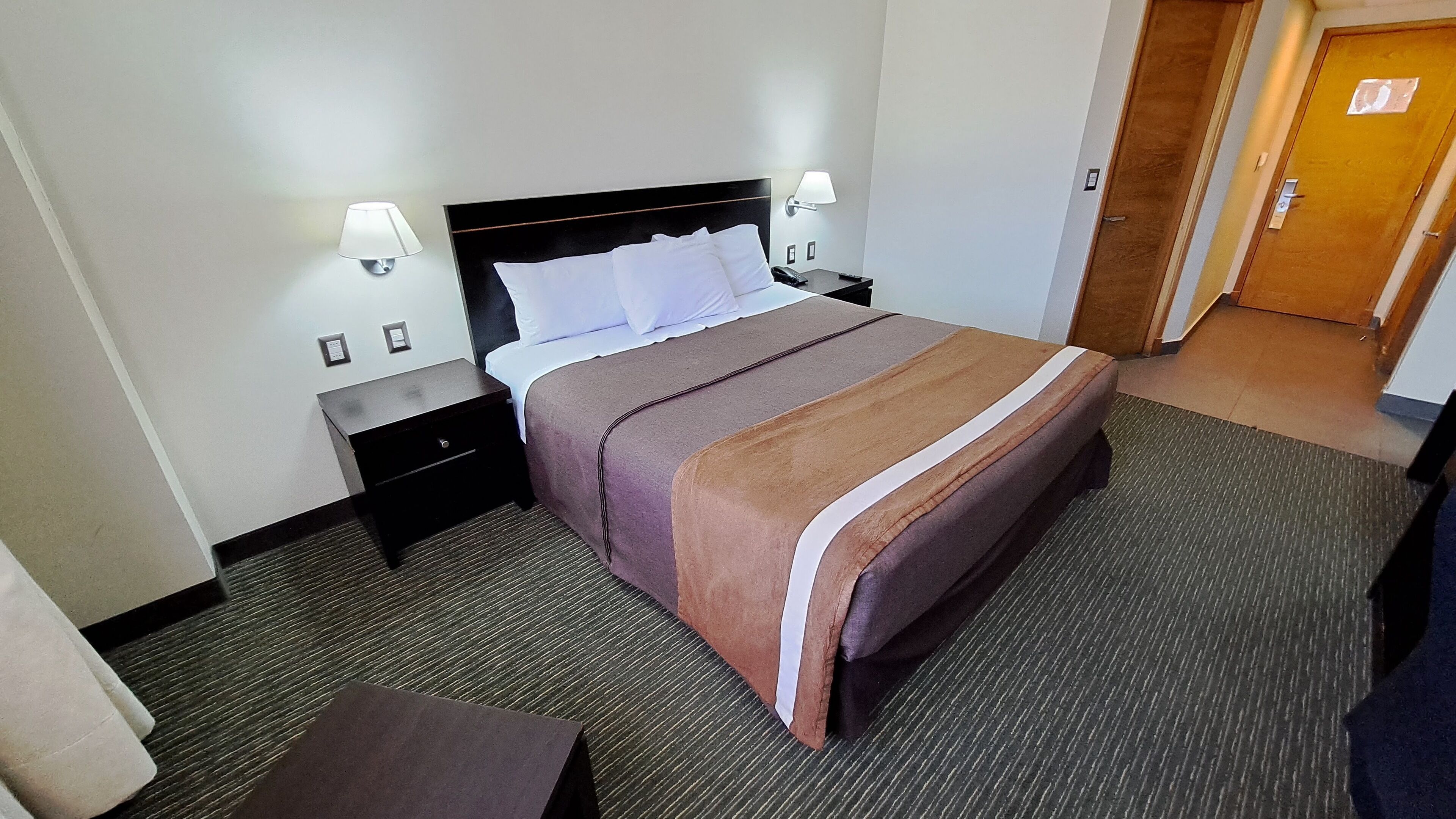 Una habitación de hotel con una cama, dos mesitas de noche, una lámpara y una puerta.