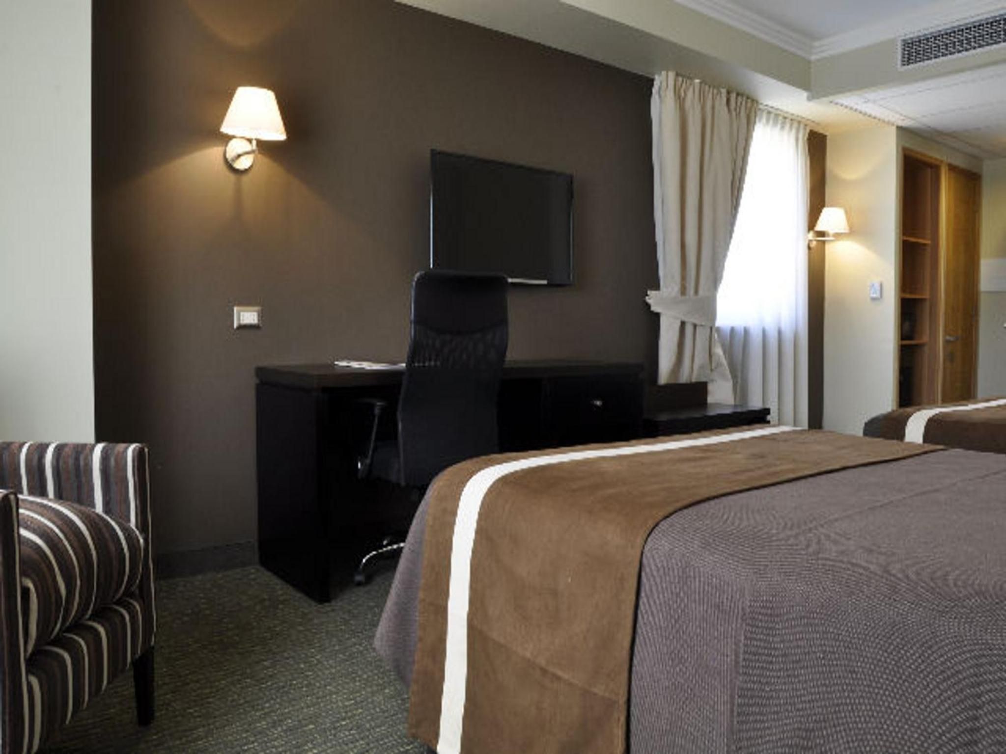 Habitación de hotel con cama, escritorio, silla, televisor y ventana con cortinas.
