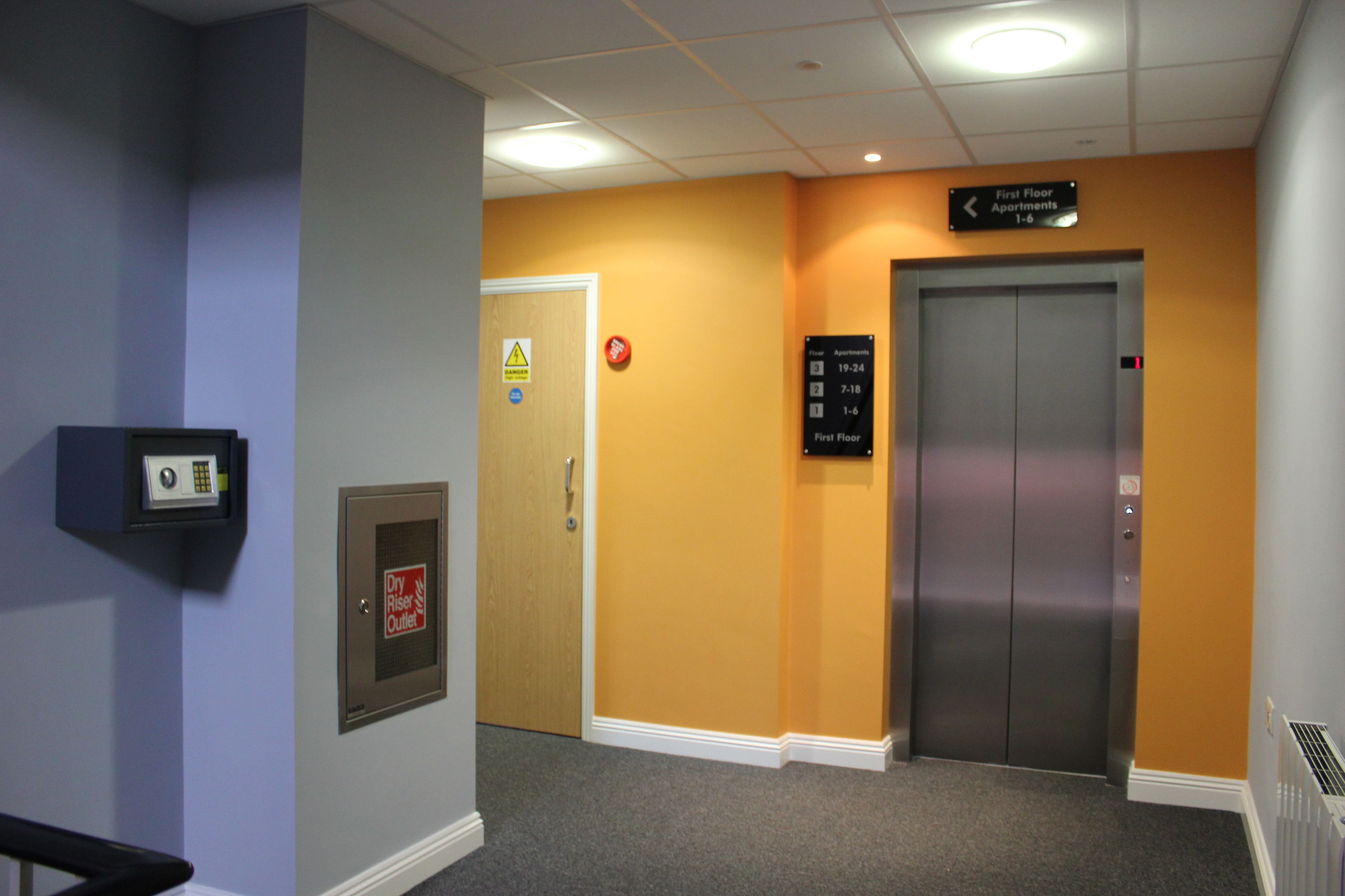 A hallway with a sign indicating first-floor apartments 1-6, an elevator, a fire alarm, and a thermostat.