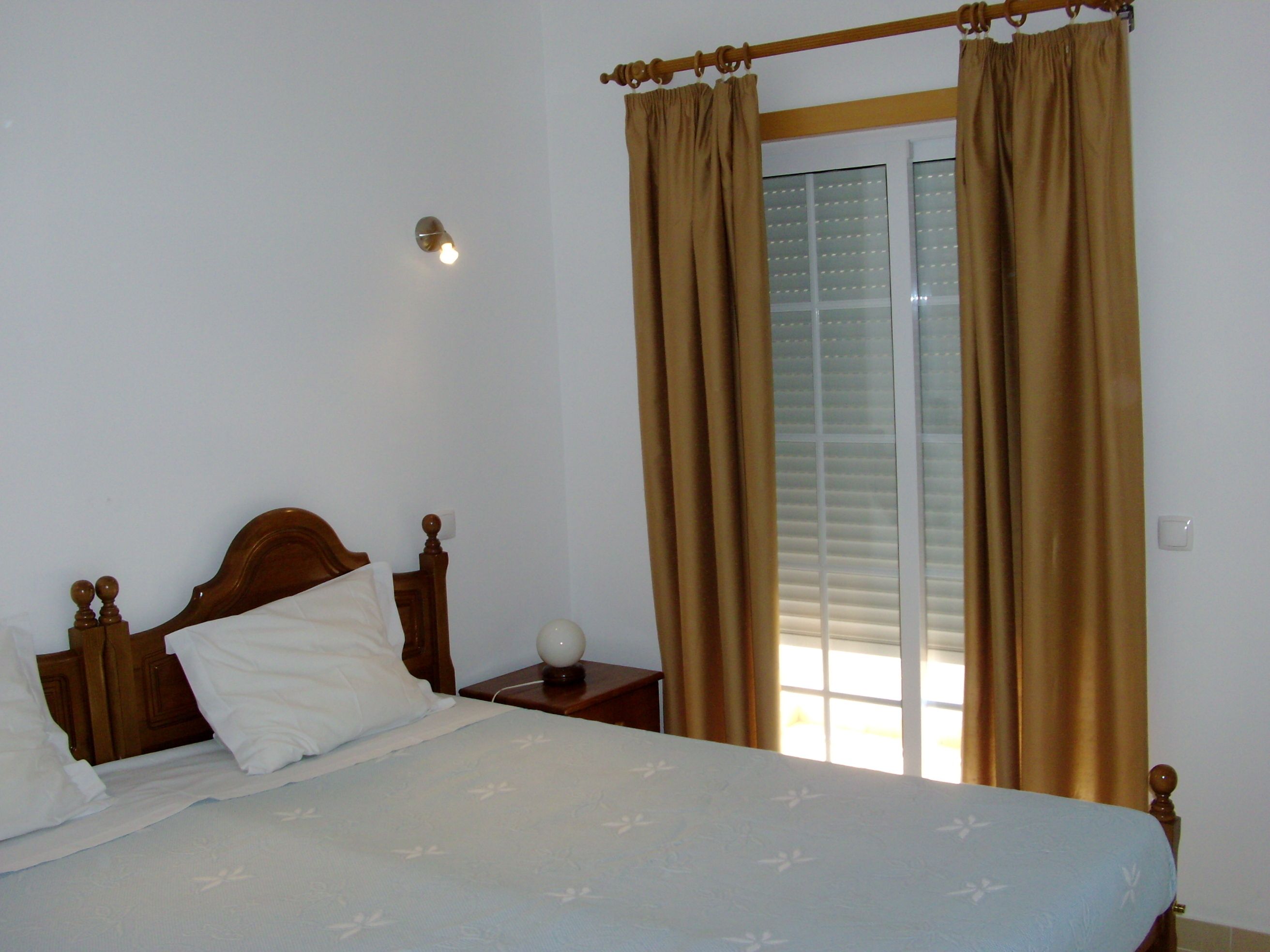 Un dormitorio con estructura de cama de madera, una mesita de noche con lámpara y una ventana con cortinas.