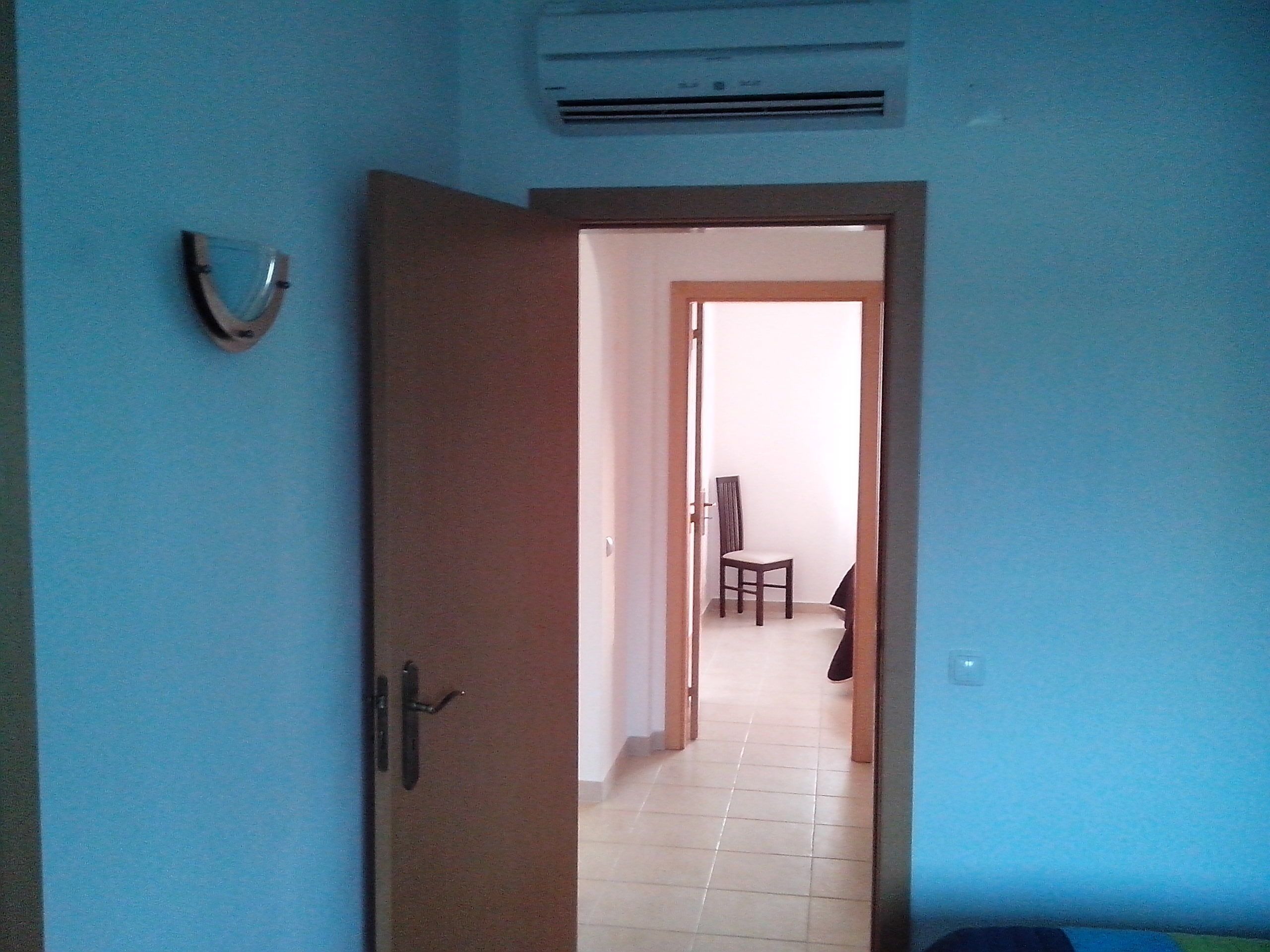 A room with a door leading to a hallway. There is a chair in the hallway and an air conditioning unit on the wall.