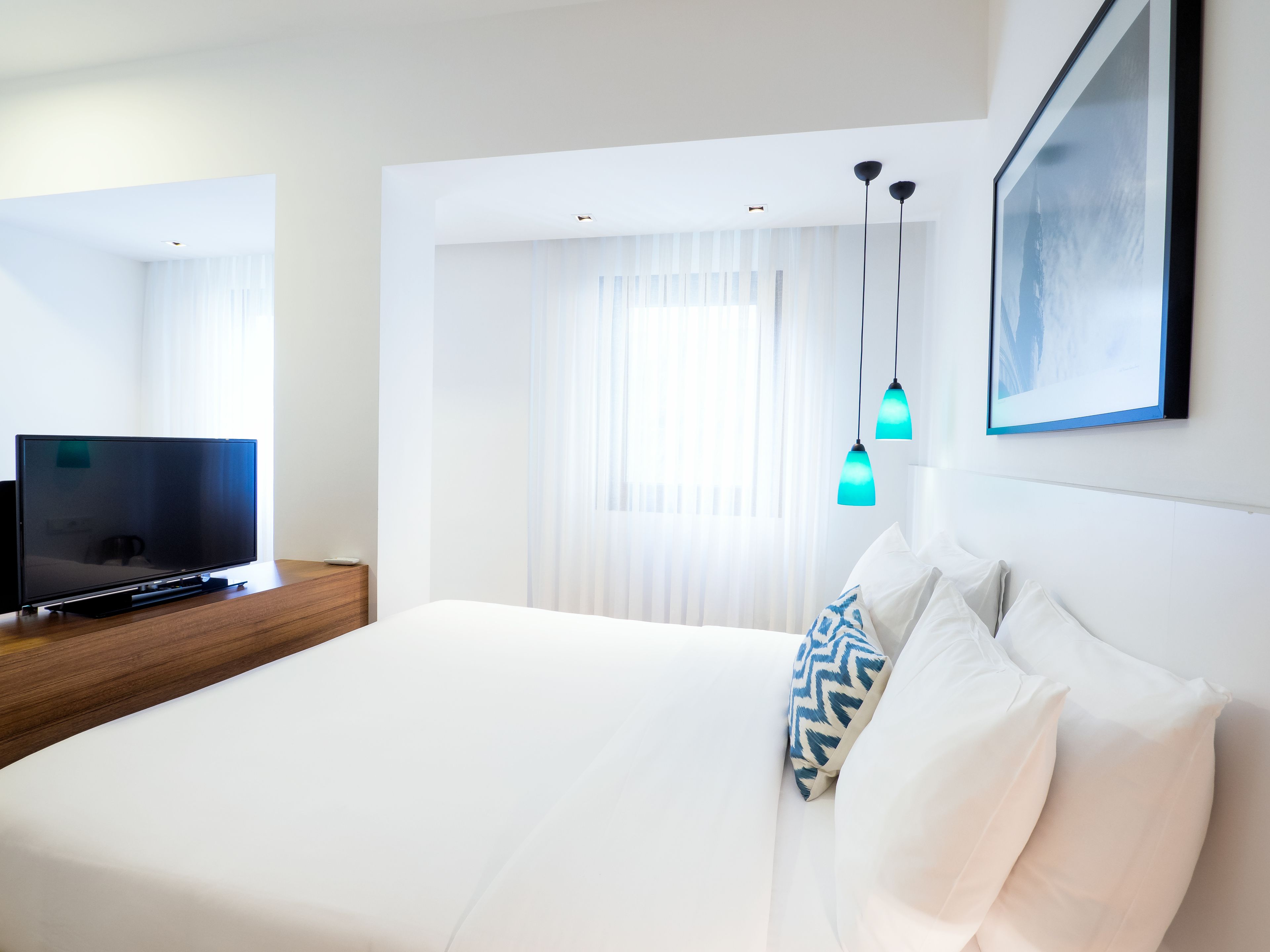 Habitación de hotel moderna con una cama grande, un televisor de pantalla plana sobre un aparador de madera y un ventanal con cortinas translúcidas.