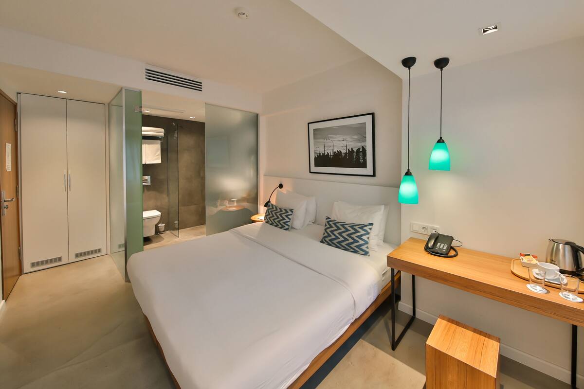 A modern hotel room with a large bed, a desk with a phone and a kettle, and a shower area.