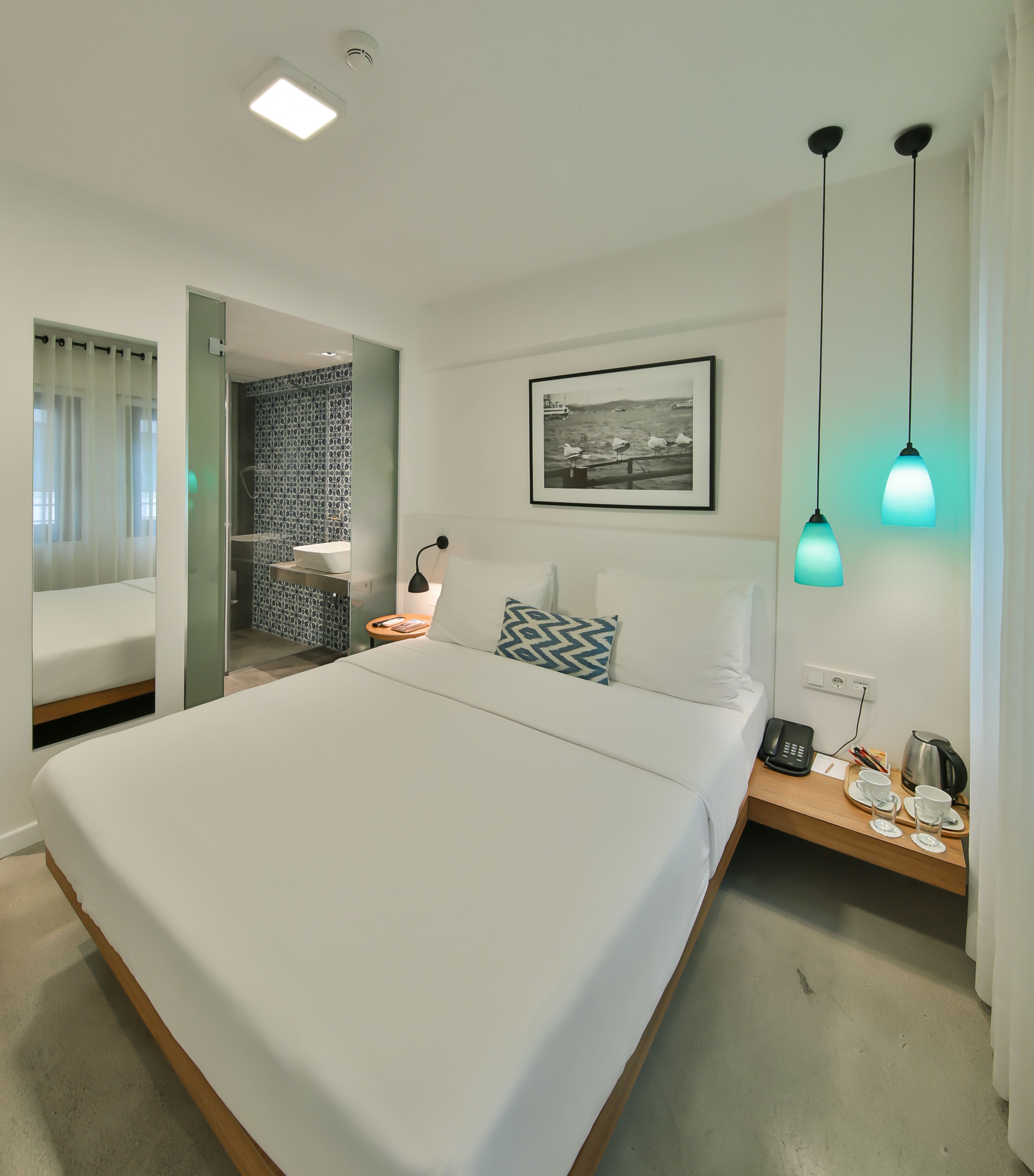 Une chambre d’hôtel moderne avec un grand lit, une table de chevet avec un téléphone et une bouilloire, une petite salle de bain avec douche et un tableau encadré au mur.