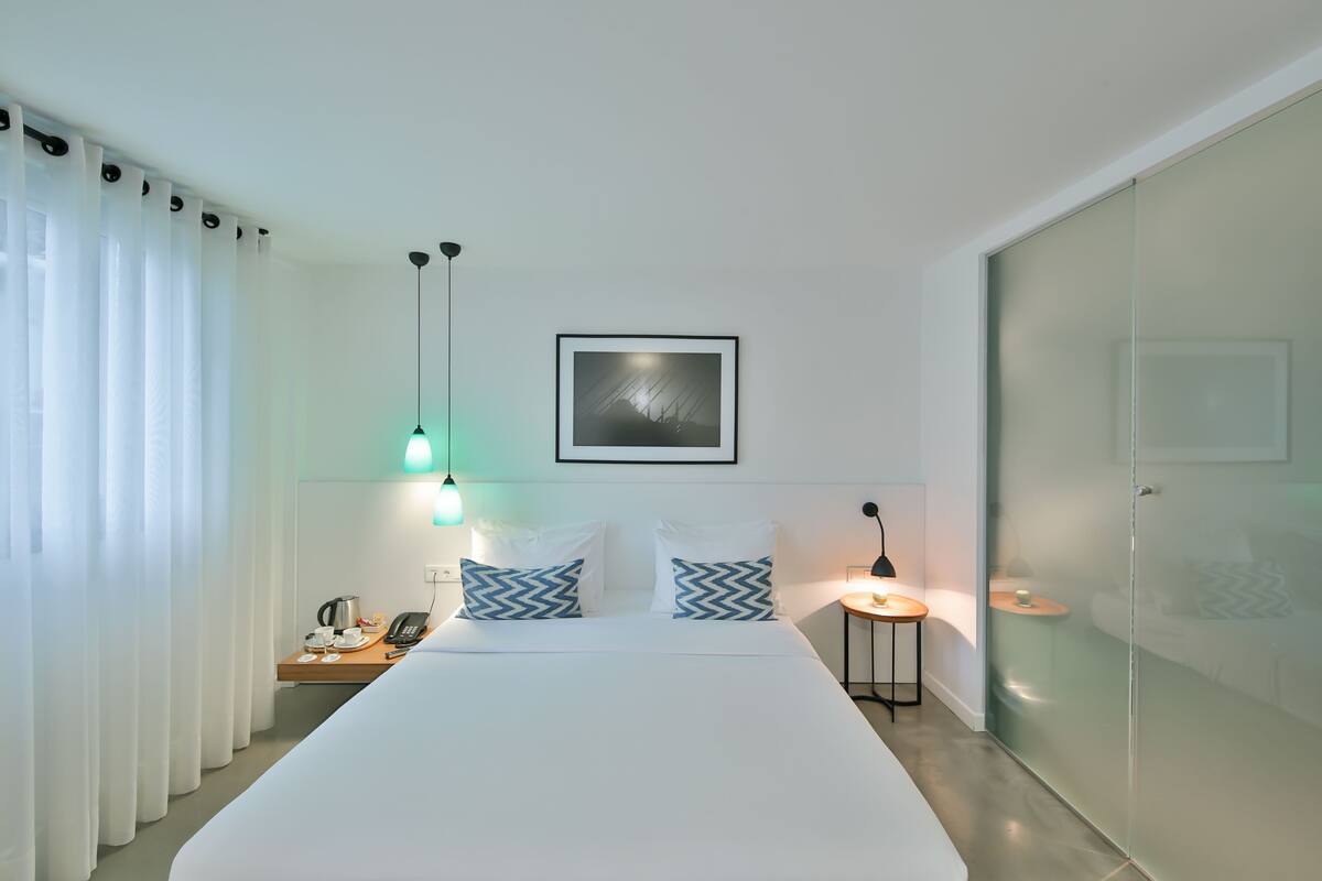 A modern hotel room with a large bed, a bedside table with a lamp, a phone, and a coffee maker. There is a framed picture on the wall above the bed.
