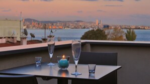 Ein Tisch mit zwei Gläsern und einer Kerze auf einem Balkon mit Blick auf eine Stadtlandschaft bei Sonnenuntergang.