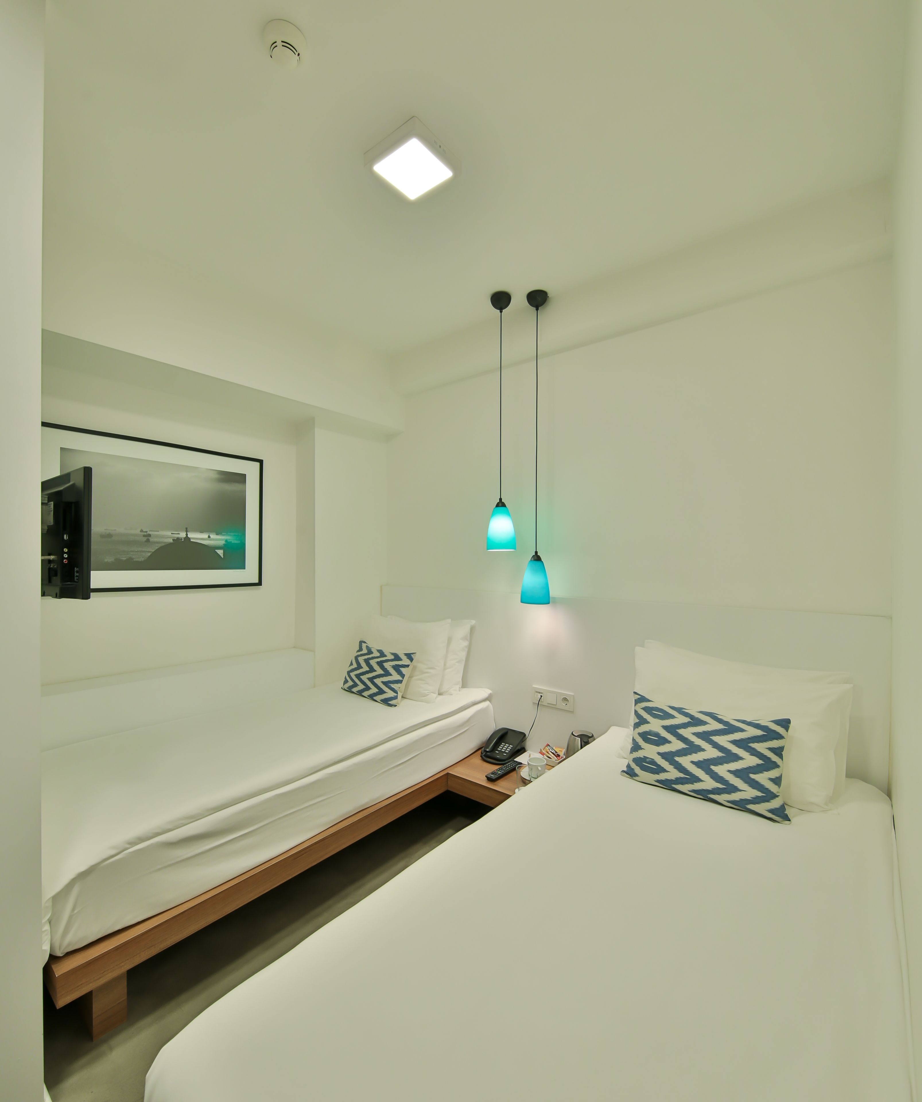 A modern hotel room with a bed, a small bedside table, a remote control, a phone, and a wall-mounted picture.