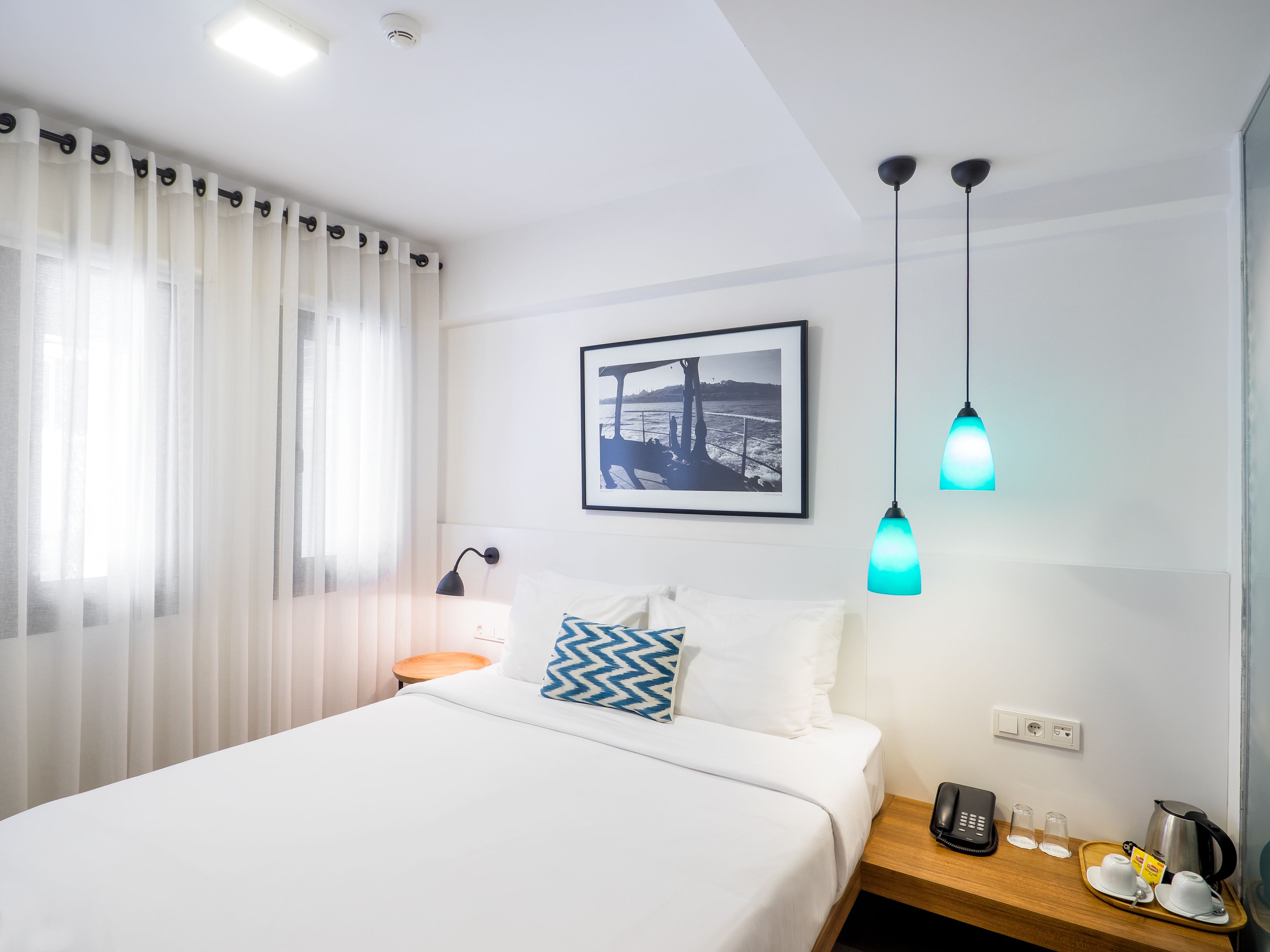 Ein Hotelzimmer mit einem Bett, einem Nachttisch mit Telefon und Brille, einem Bild an der Wand und einem Fenster mit Vorhängen.