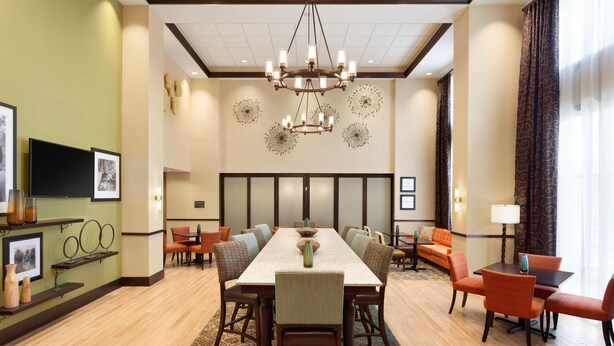 Uma sala de jantar de hotel com uma mesa comprida, várias cadeiras, um candelabro e uma televisão na parede.