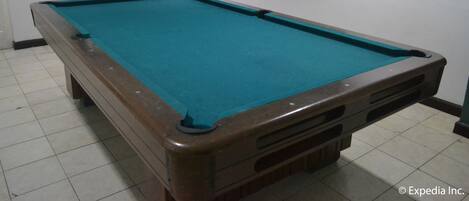 A pool table with a green felt surface, a wooden border, and a corner pocket. There is a framed picture on the wall above the table and a mirror on the wall to the left.