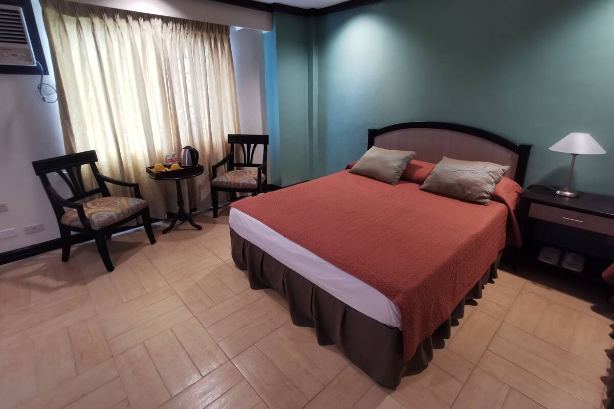 Quarto de hotel com cama grande, escrivaninha, duas cadeiras e janela com cortinas.