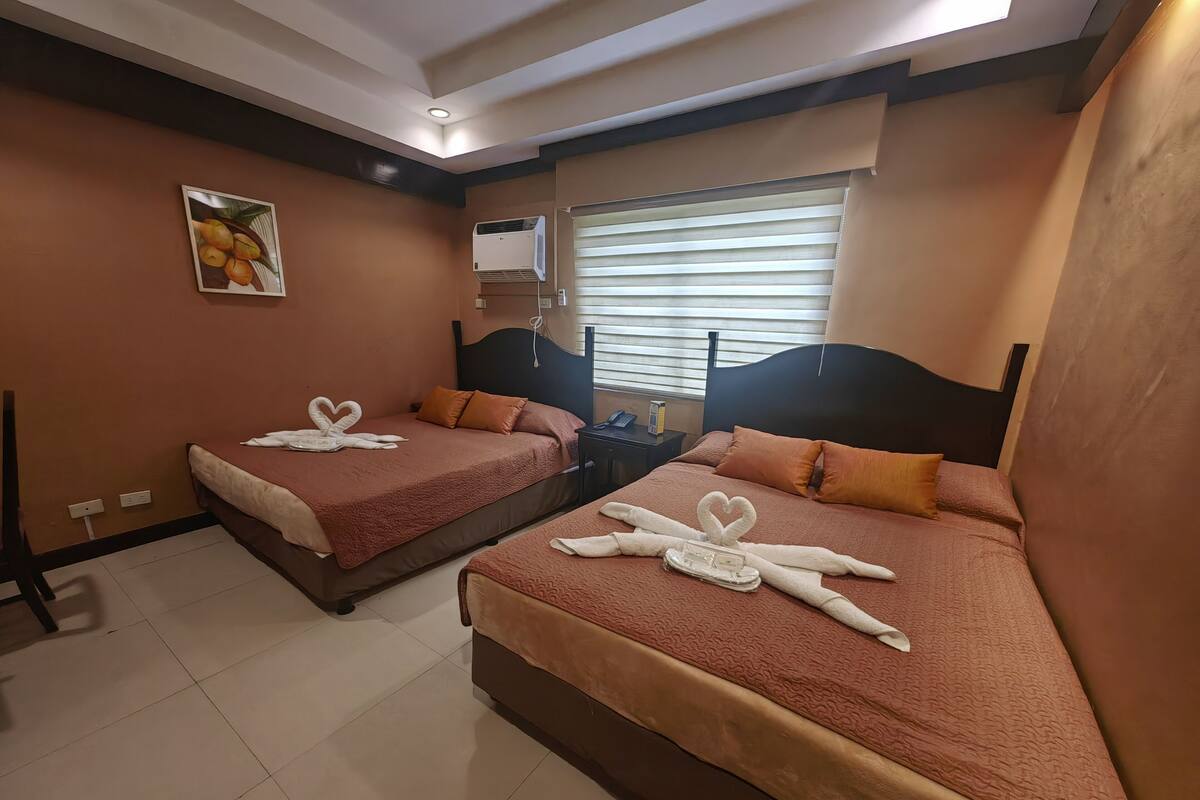 Quarto de hotel com duas camas, cada uma decorada com toalhas em formato de coração, um ventilador de teto, ar-condicionado fixado na parede e uma janela com persianas.
