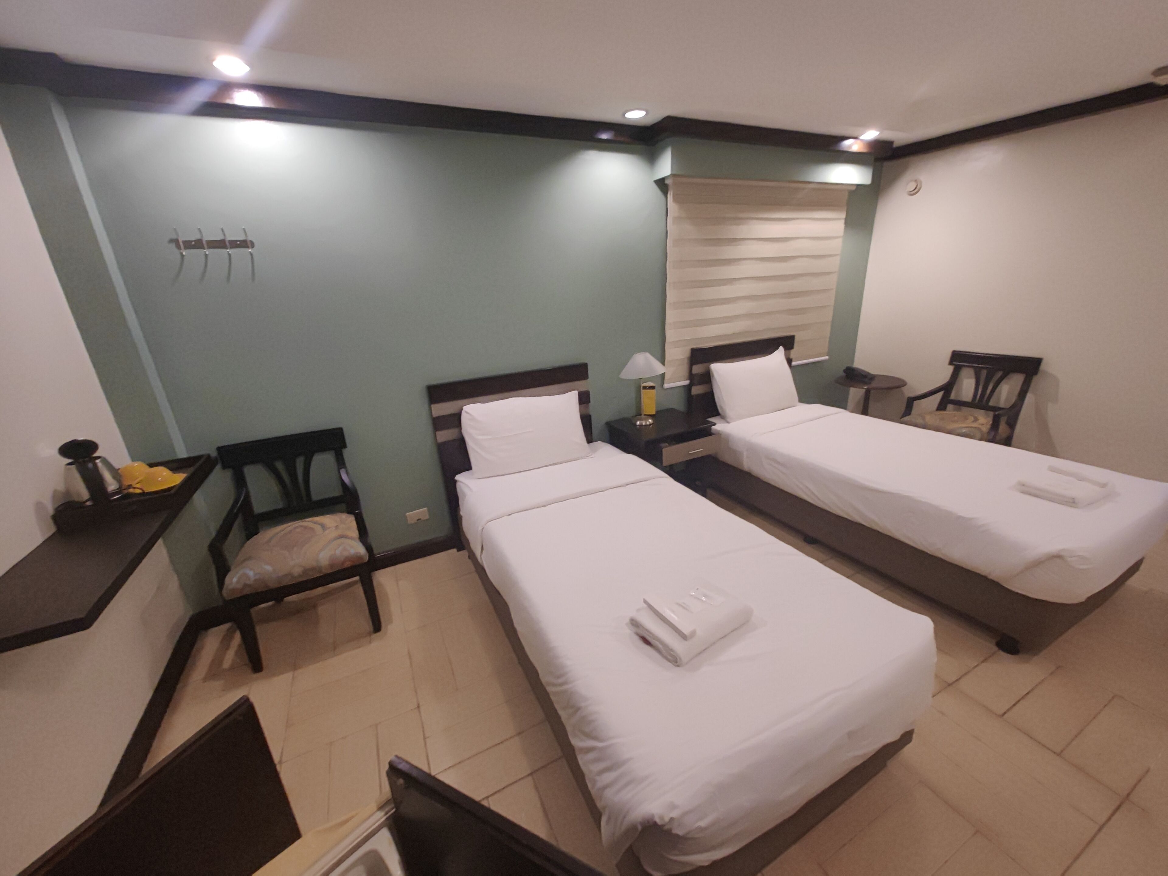 A hotel room with two single beds, a small desk, a chair, a window with blinds, and a wall-mounted hook.