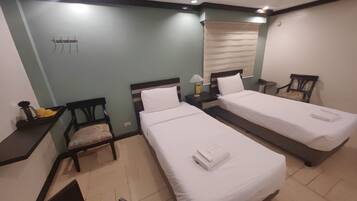 A hotel room with two single beds, a small desk, a chair, a window with blinds, and a wall-mounted hook.
