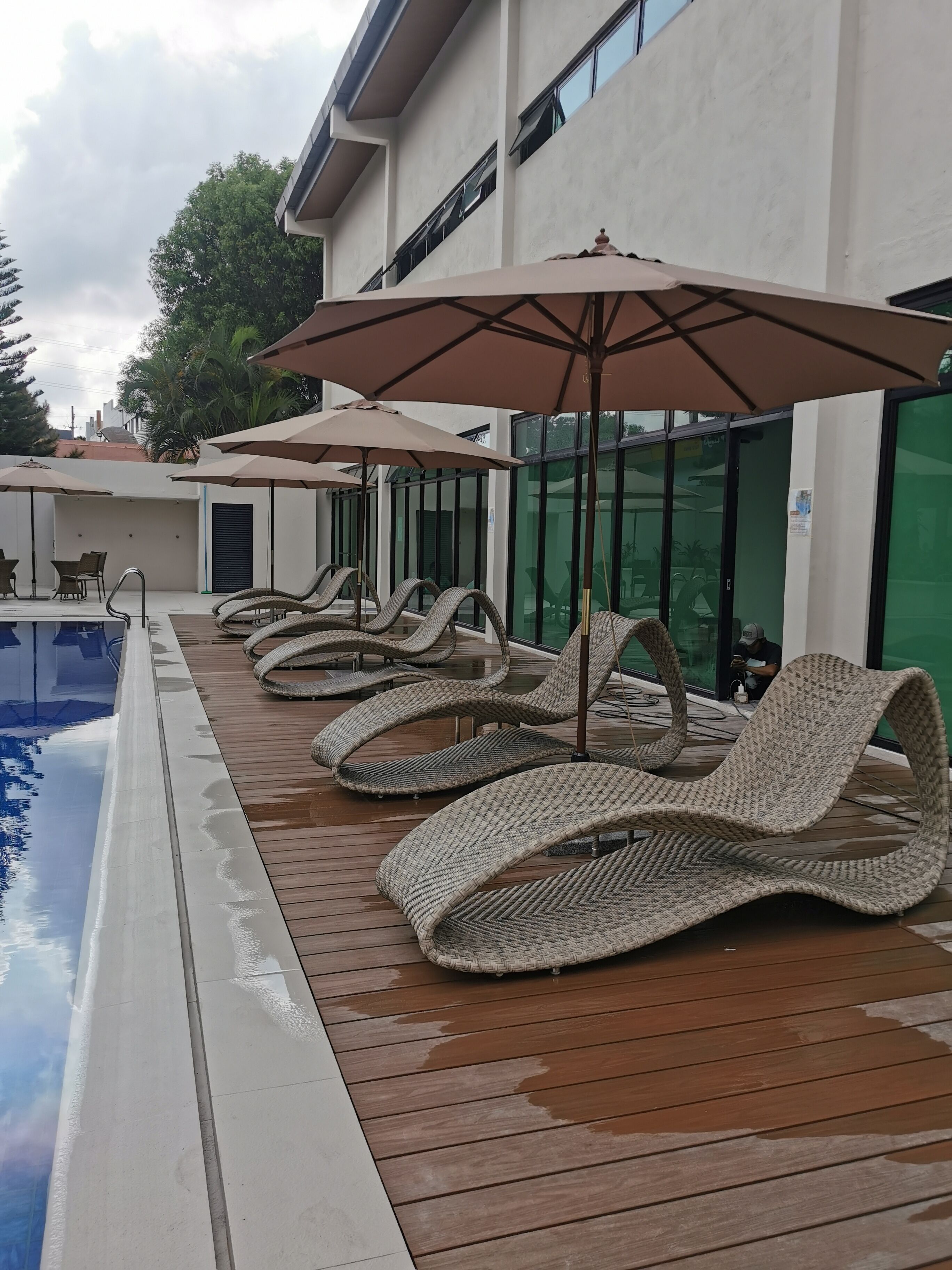 A poolside area with lounge chairs and umbrellas, a wooden deck, and a glass-enclosed building.