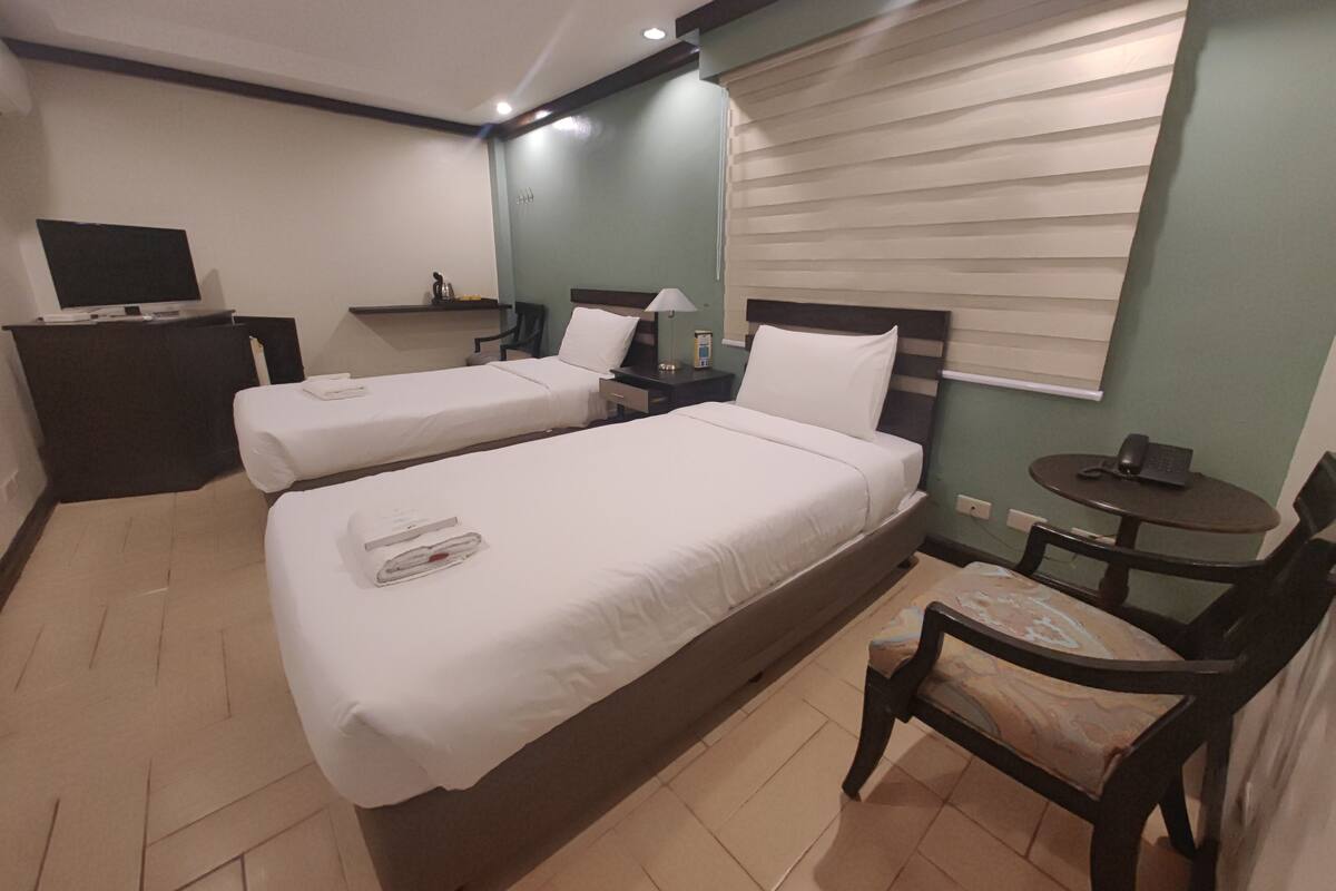 Quarto de hotel com duas camas, uma escrivaninha, uma cadeira, televisão e janela com persianas.