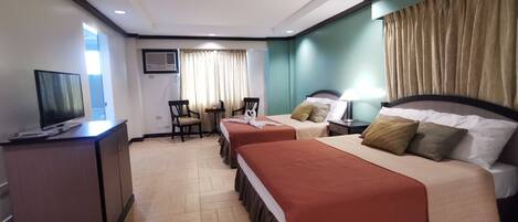 Habitación de hotel con dos camas, un escritorio con televisor, una pequeña mesa con sillas y un ventanal con cortinas.