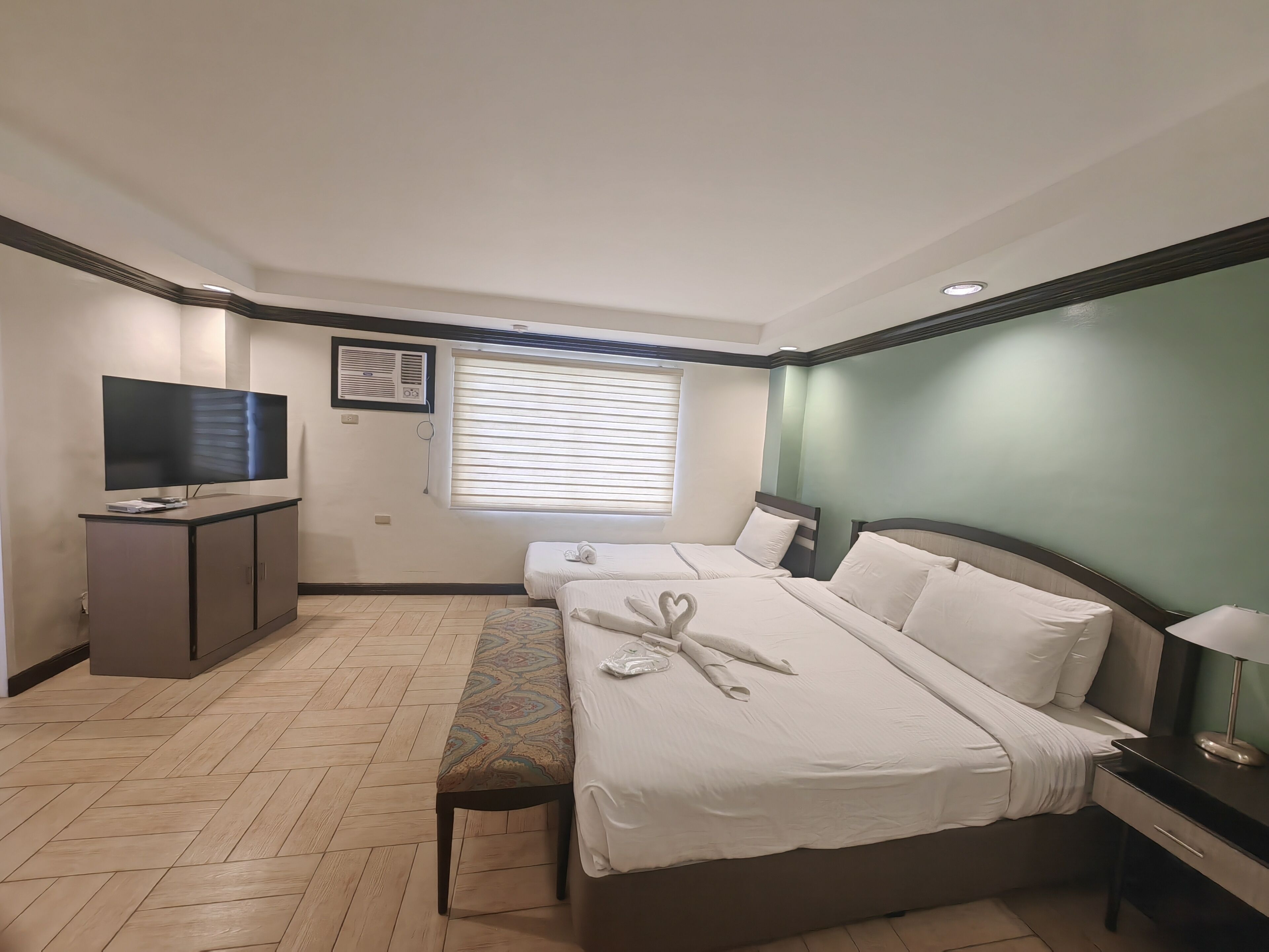 A hotel room with two beds, a TV, a bench, and a window with blinds.