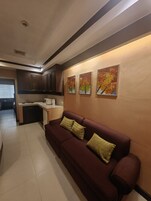 Una sala de estar con un sofá de color oscuro, una cocina con armarios y un microondas, y tres cuadros en la pared.